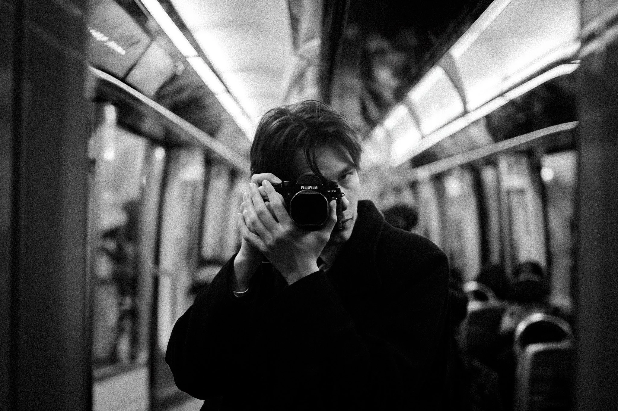 Un jeune homme avec une veste noire, tenant un appareil photo Fujifilm, se prenant en selfie dans un train en mouvement, en noir et blanc.