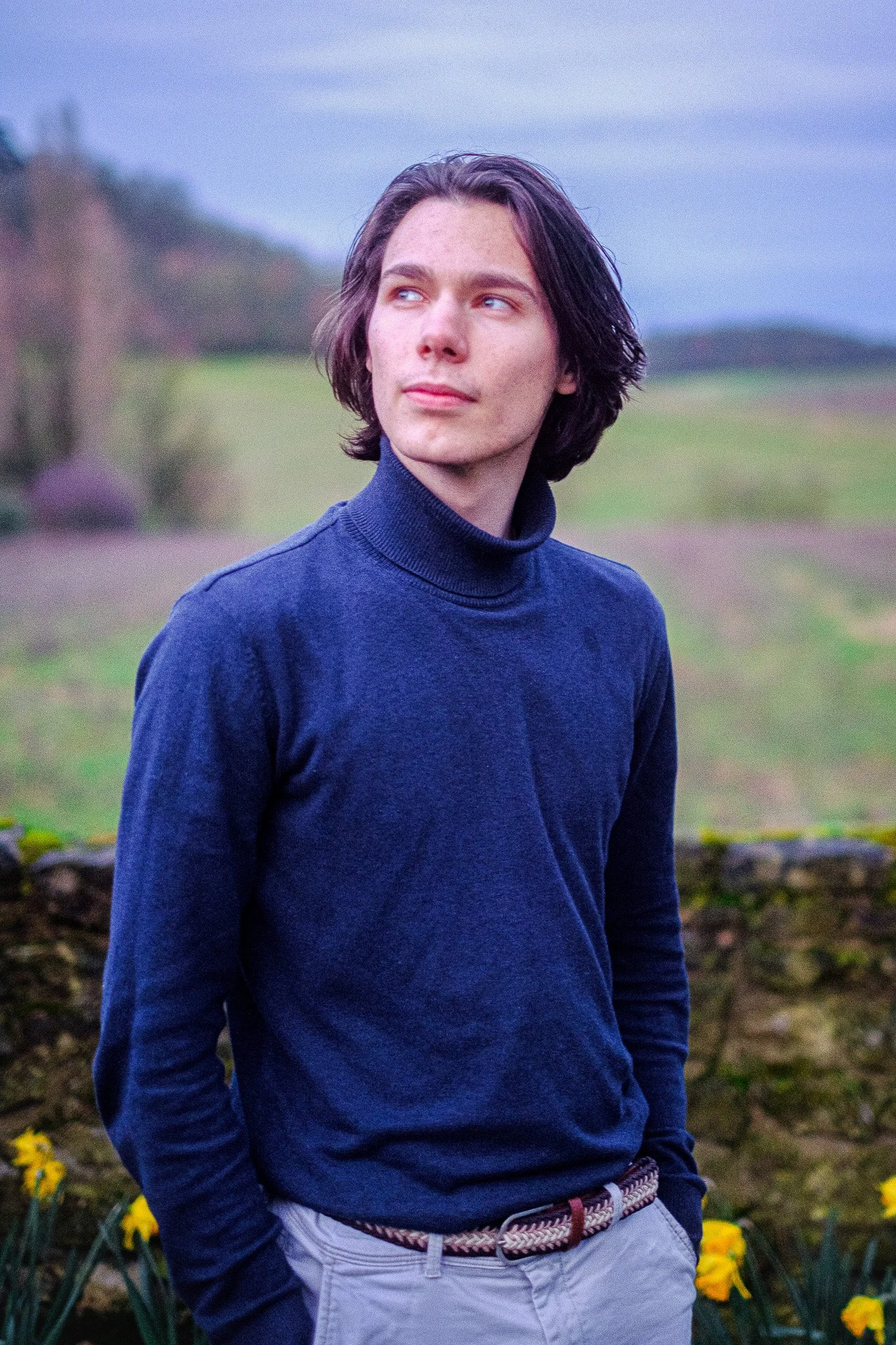 Jeune homme aux cheveux longs, portant un pull bleu marine et un pantalon beige, se tenant en plein air avec un paysage de champs et de collines en arrière-plan.