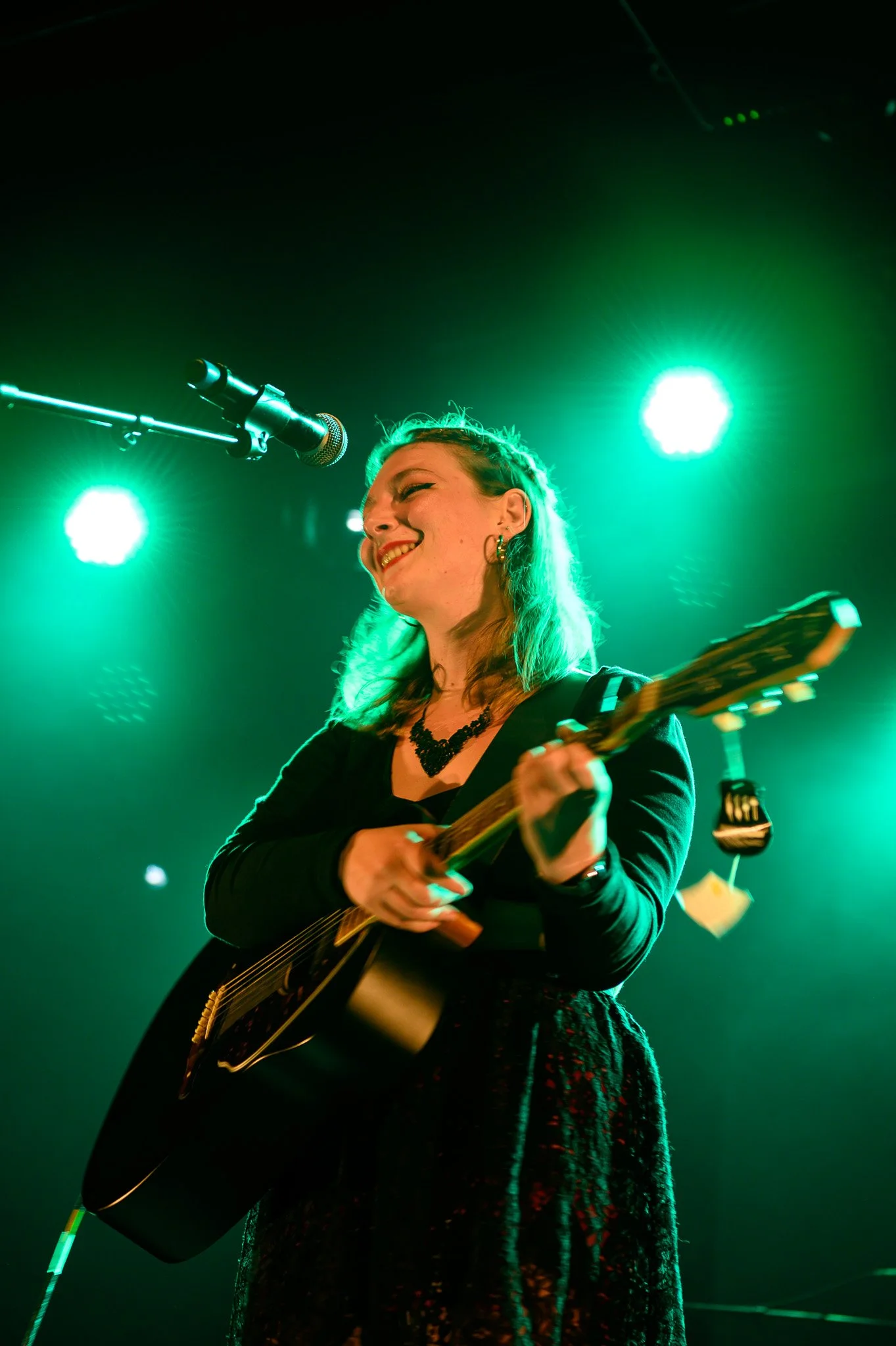 Une femme souriante, jouant de la guitare électrique, se produisant sur scène avec une lumière verte en arrière-plan.