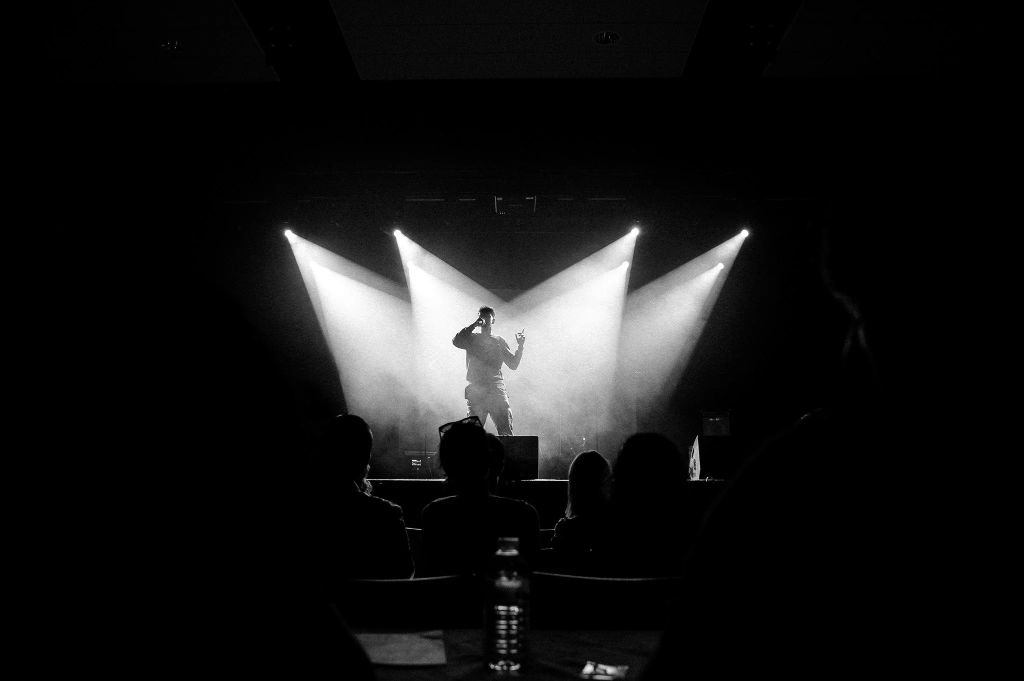 Une scène de spectacle avec un artiste sur scène, éclairé par des projecteurs, dans un environnement noir et blanc.