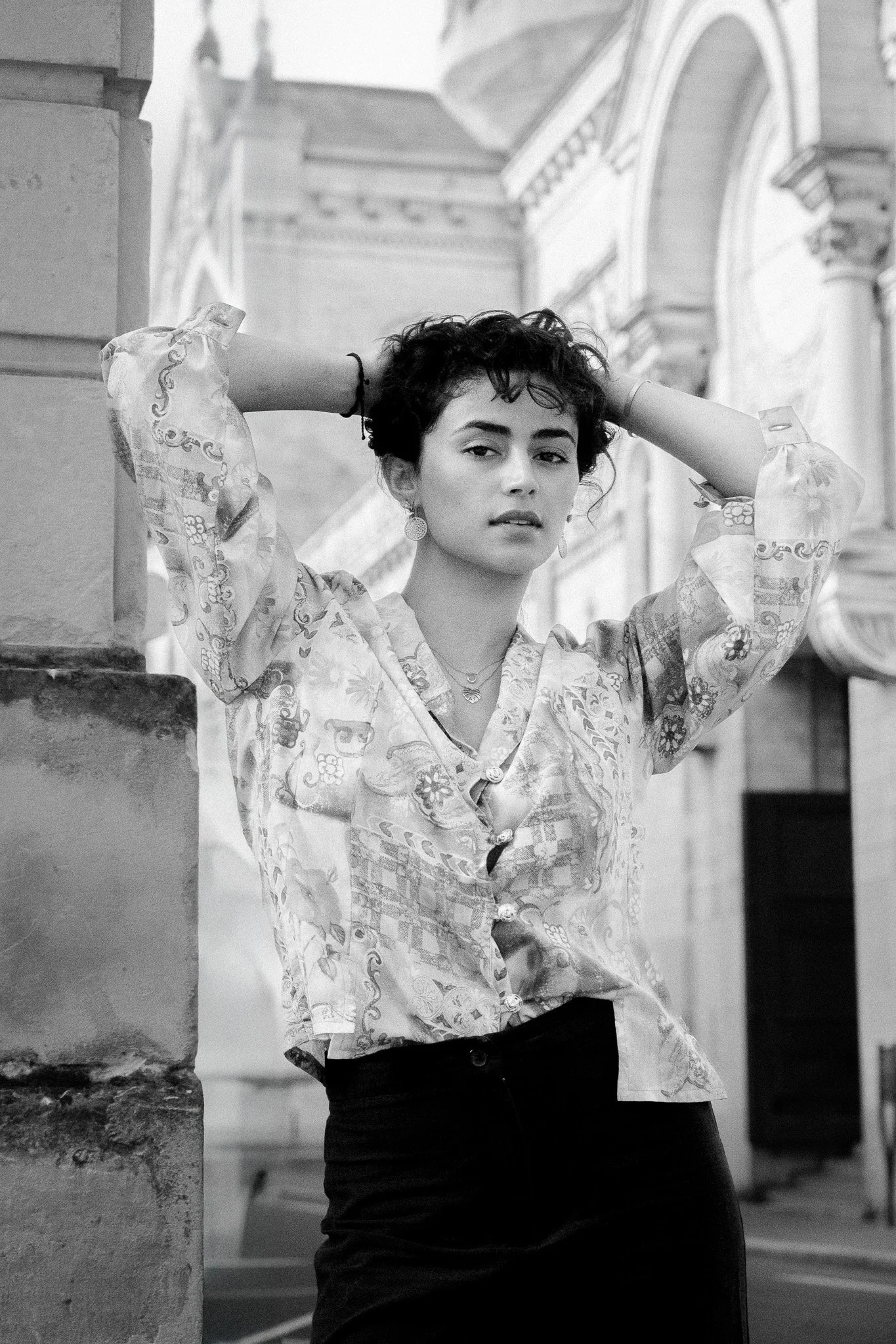 Une jeune femme posant avec les bras levés derrière la tête dans une rue architecturale, en noir et blanc.