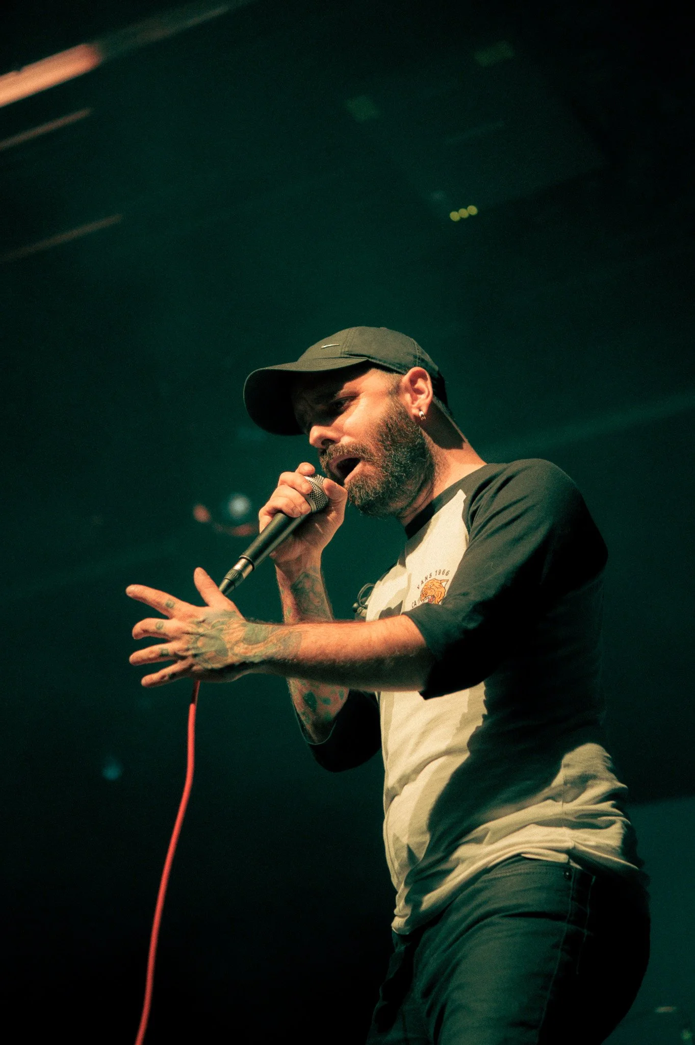 Un homme musculaire porte un chapeau de baseball, une moustache et une barbe, chante dans un micro lors d'une performance sur scène, avec un éclairage tamisé en arrière-plan.