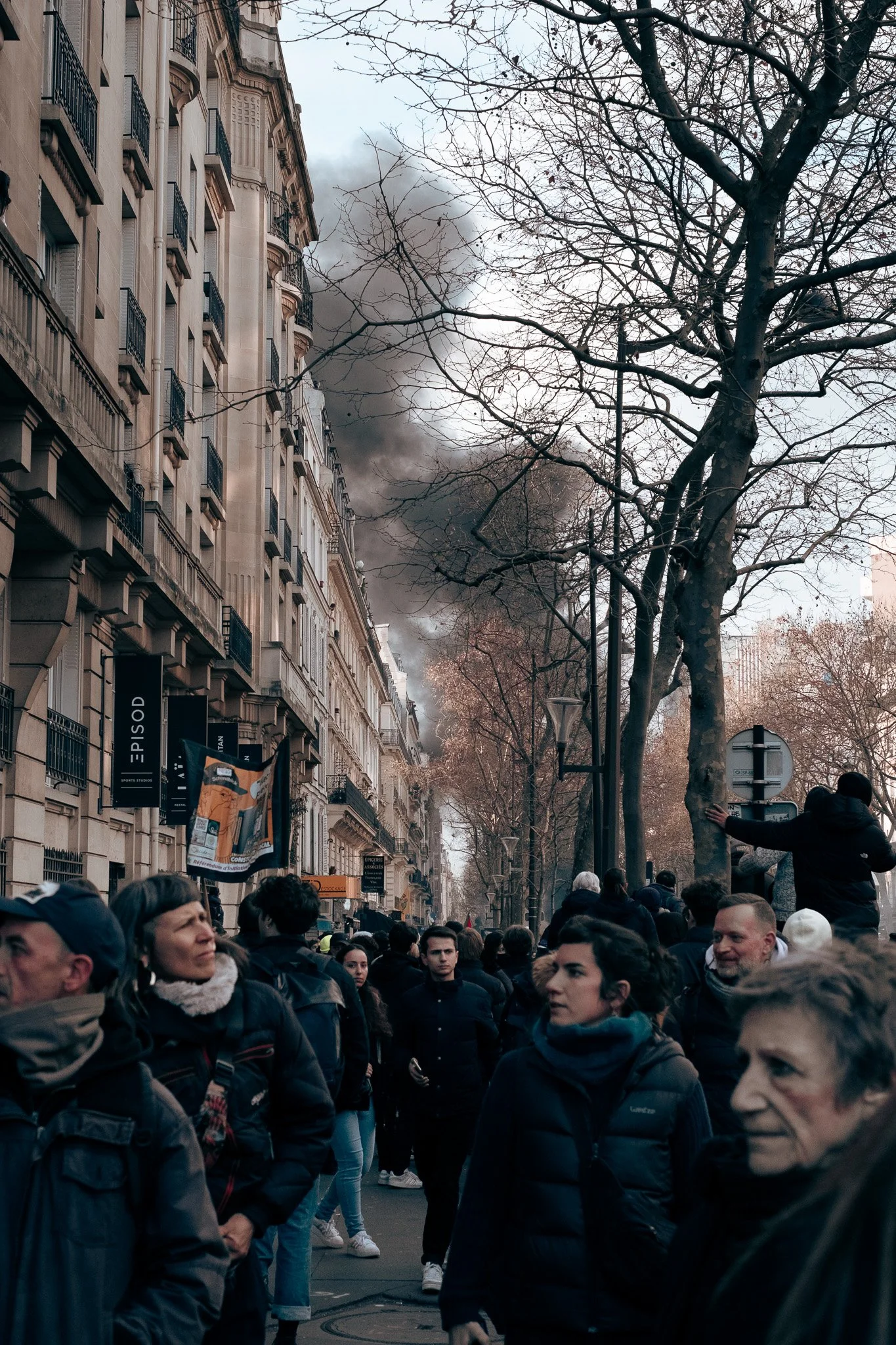 Une rue urbaine avec des bâtiments à gauche et des arbres sans feuilles à droite. Il y a un groupe de personnes, certains regardant vers le haut, tandis que de la fumée noire s'élève dans le ciel en arrière-plan.