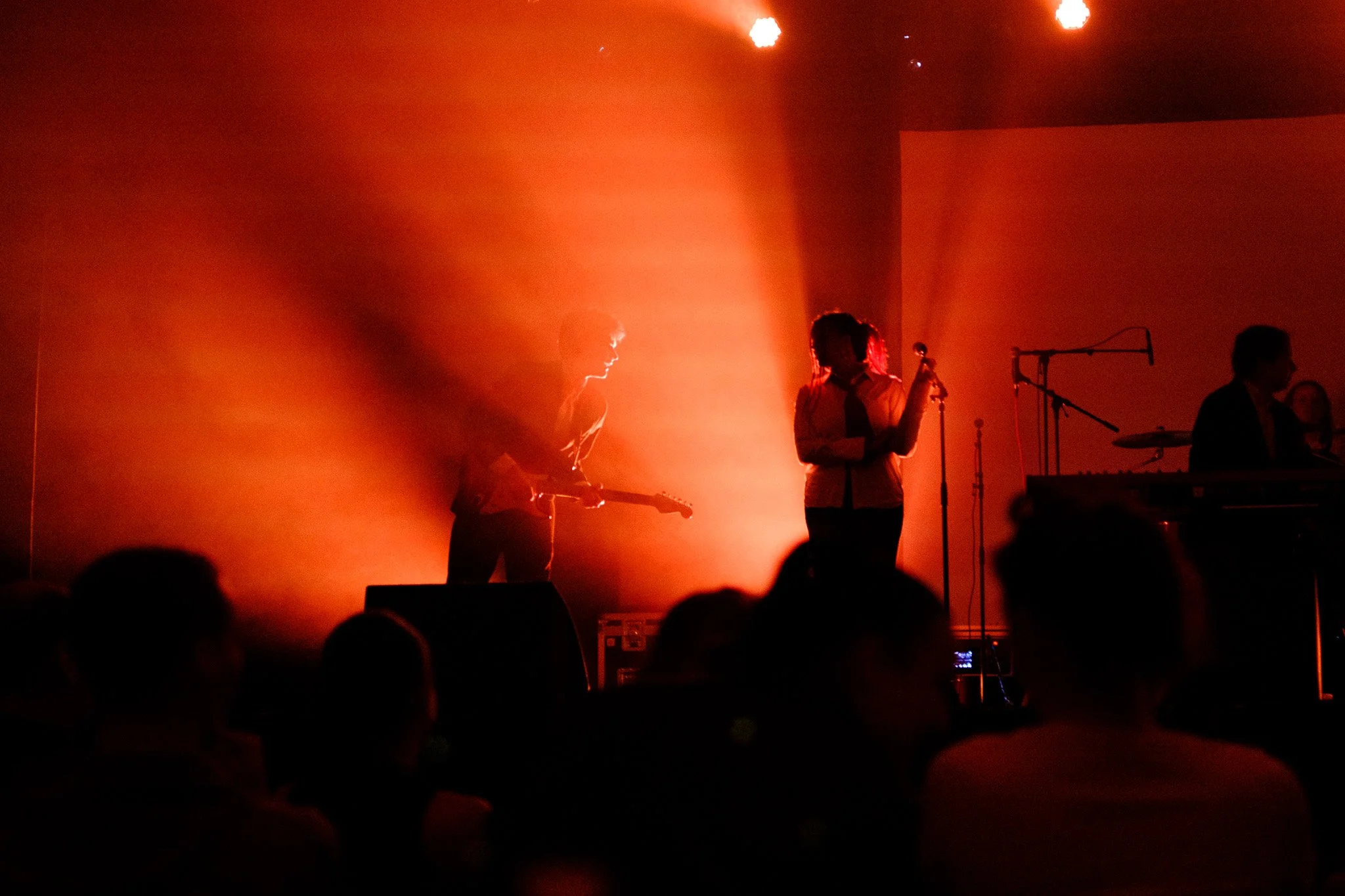 Un groupe de musiciens en performance sur scène, éclairés par une lumière orange et rouge, avec le public en premier plan.