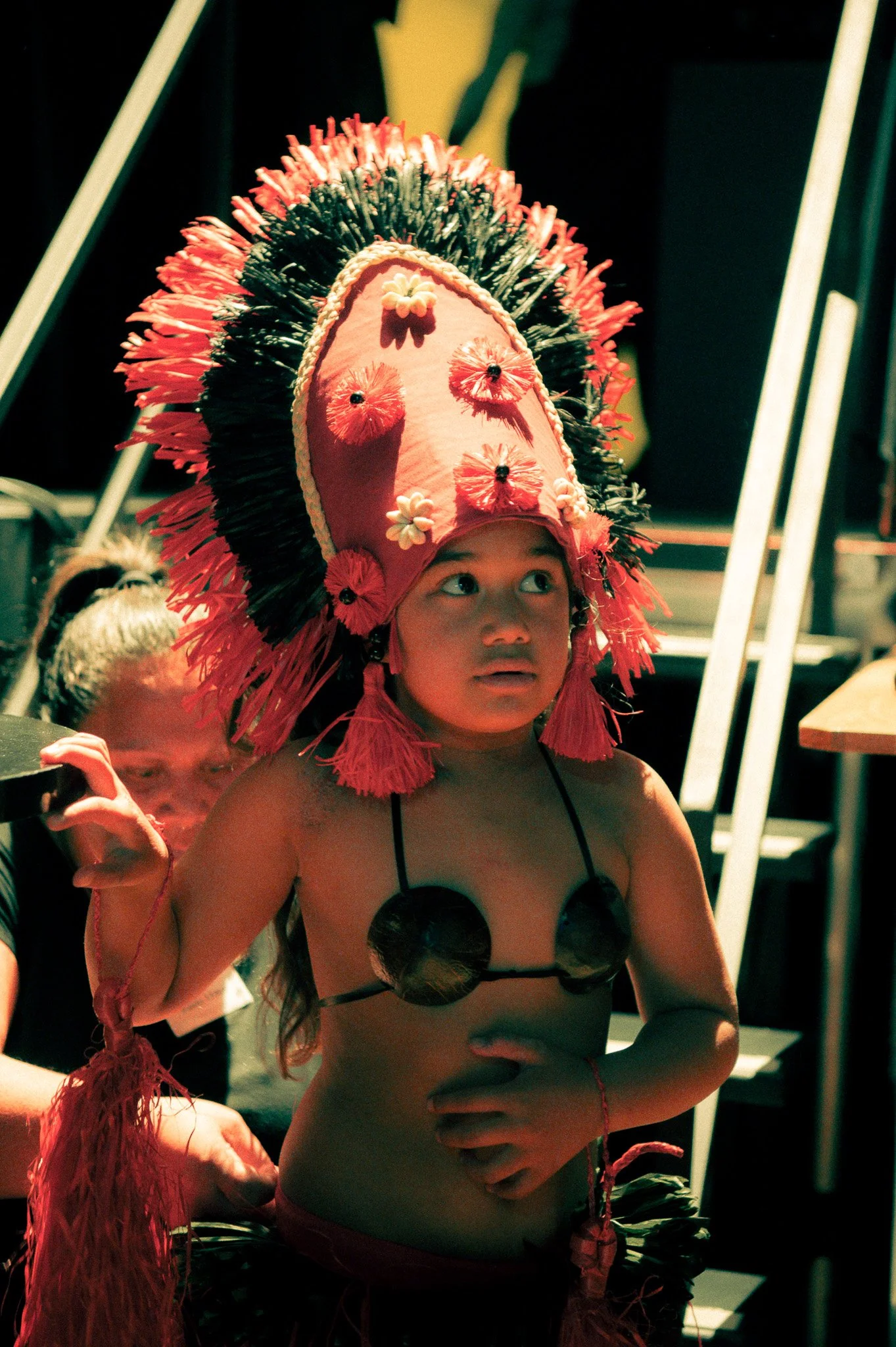 Un jeune garçon en costume traditionnel avec un grand chapeau orné de fleurs, de plumes et de décoration colorée, à l'air pensif ou surpris.