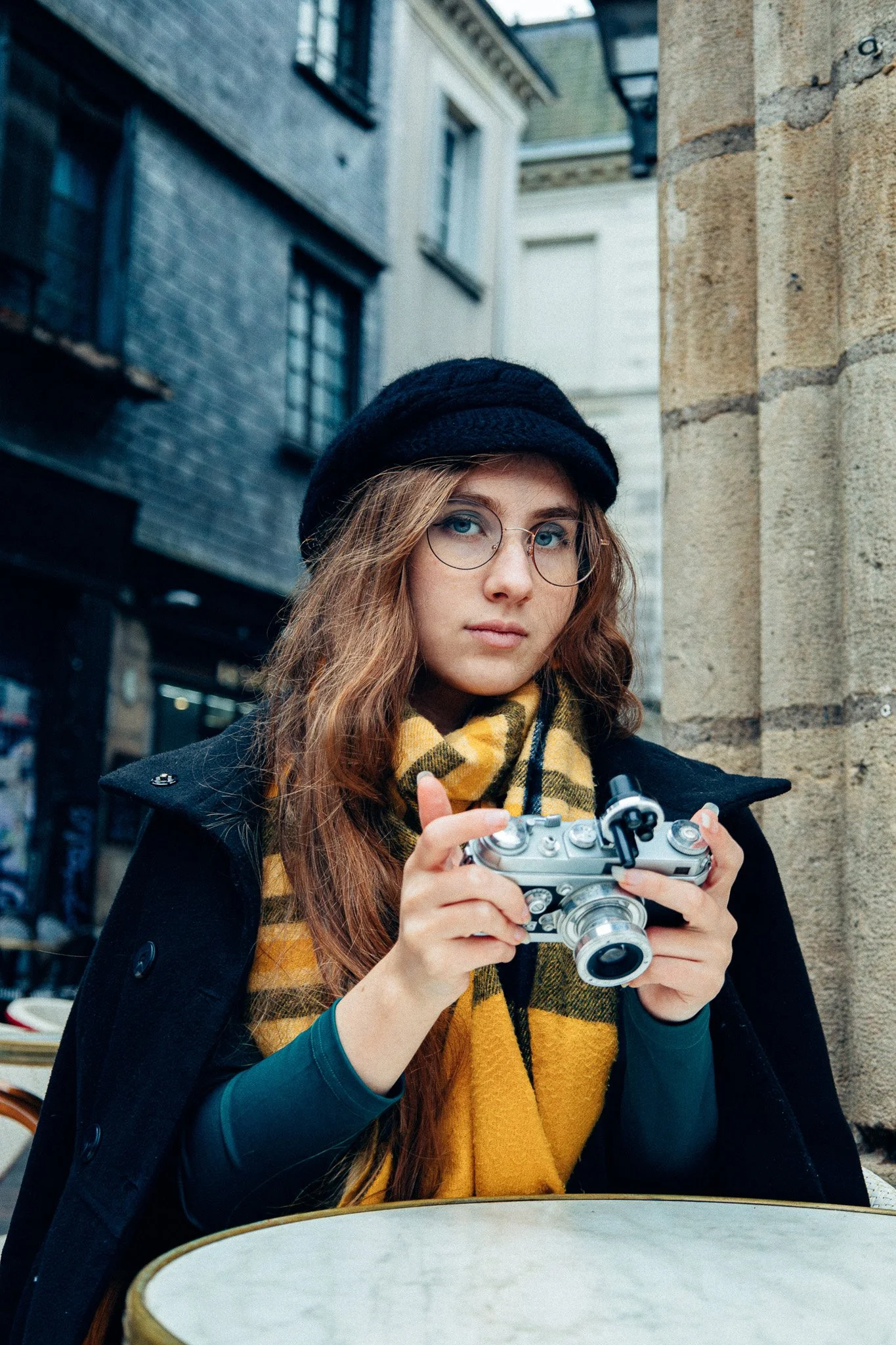 Jeune femme avec lunettes, bonnet noir et écharpe jaune à carreaux, tenant un appareil photo, assise à une table en plein air, rue urbaine