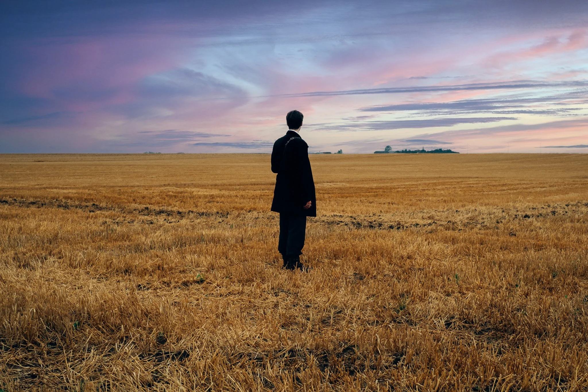 Un homme en costume noir se tient seul dans un champ doré au coucher du soleil, regardant vers l'horizon avec des nuages colorés dans le ciel.