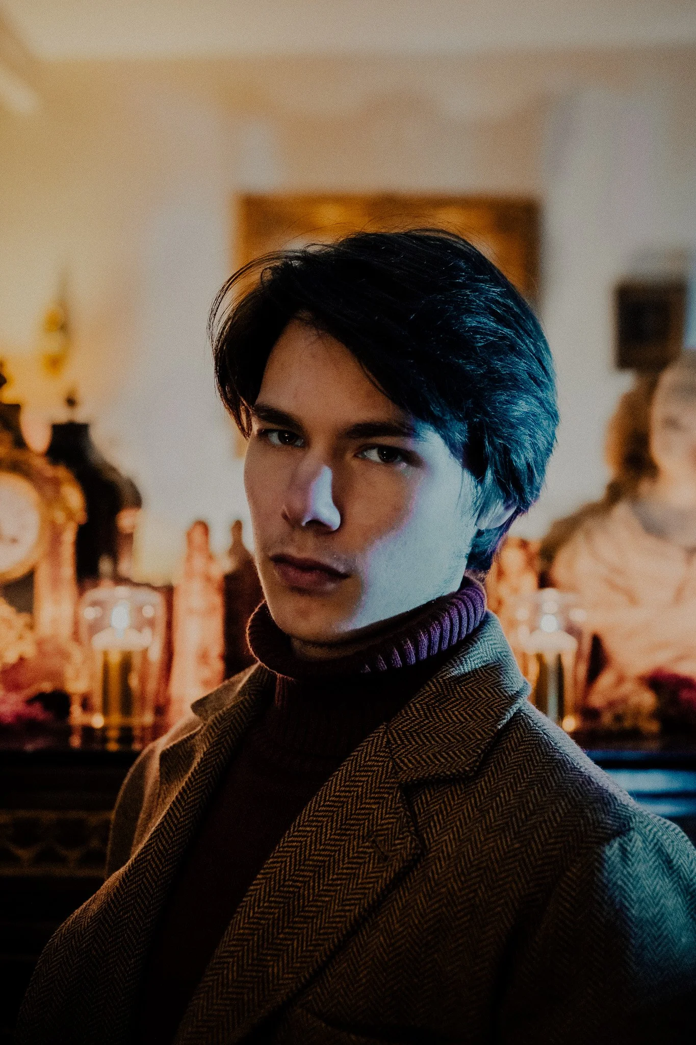 Jeune homme aux cheveux noirs, portant un manteau à carreaux et un turtleneck violet, posé dans un intérieur avec un décor ancien et une horloge en arrière-plan.
