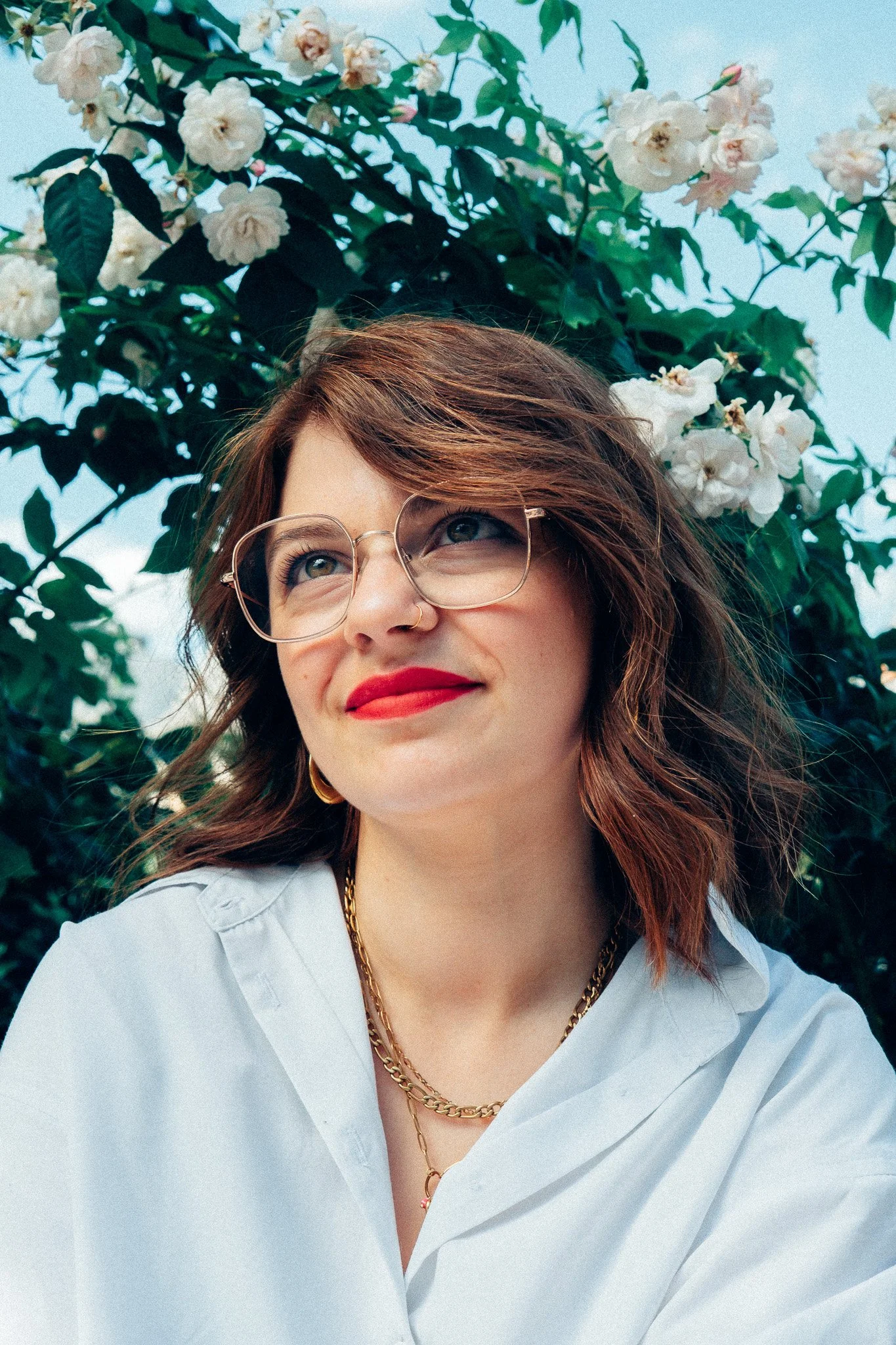 Jeune femme souriante portant des lunettes, un maillot blanc, avec des bijoux en or, debout devant un buisson de fleurs blanches et vertes.