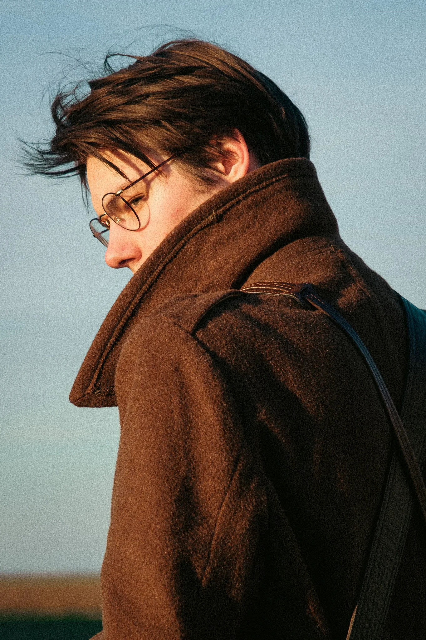 Jeune homme portant des lunettes de soleil et une veste en laine marron, vue de profil, contre un ciel clair.