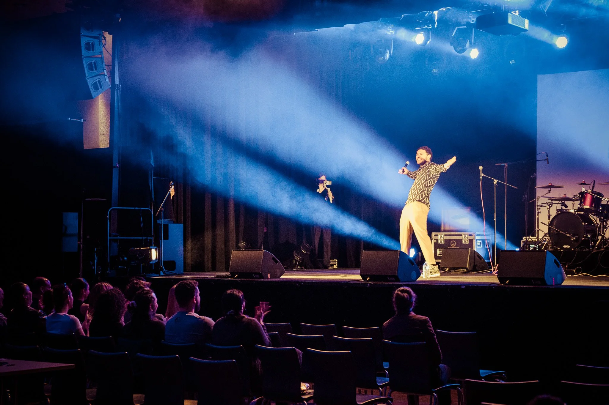 Un artiste sur scène en train de chanter et de danser avec un micro, sous des lumières bleues, devant un public assis.