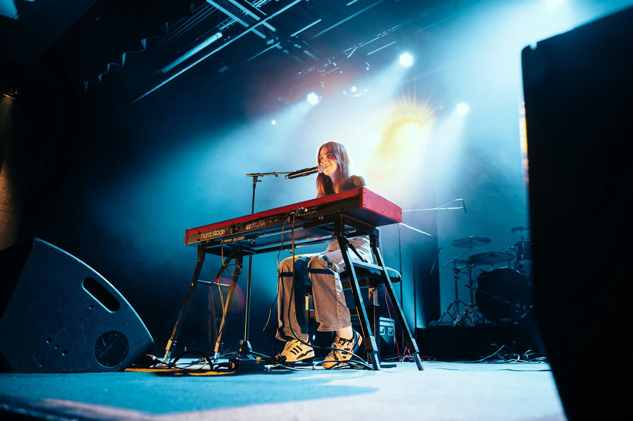 Une femme joue du clavier sur scène avec un microphone, sous des lumières colorées, dans un environnement de concert.