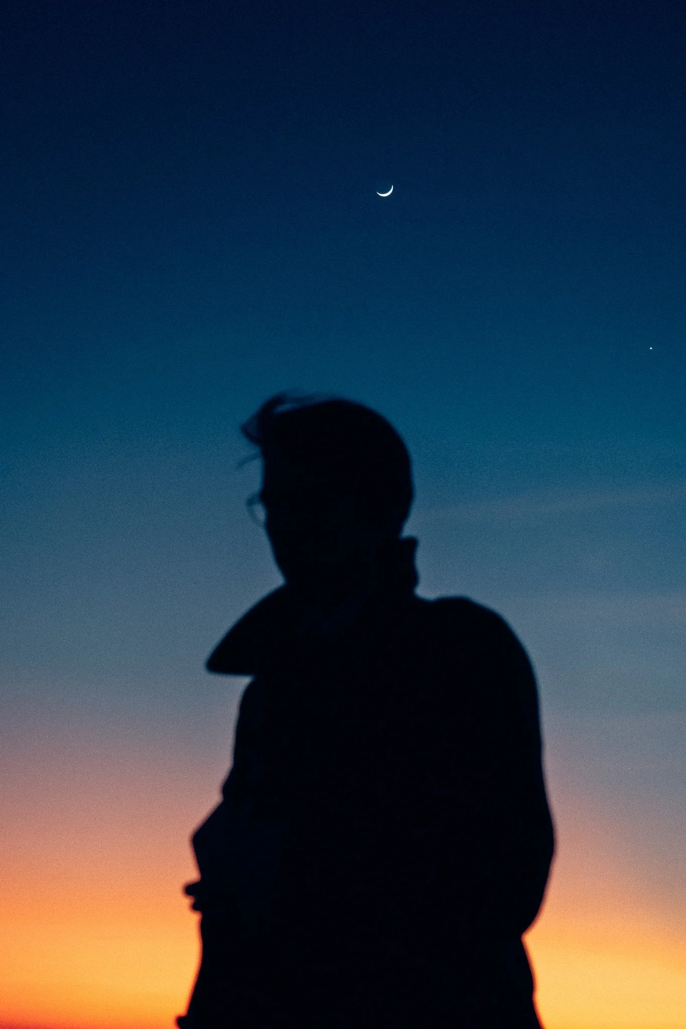 Silhouette d'un homme avec un ciel étoilé derrière, comprenant la lune en croissant et une étoile, lors du coucher du soleil.