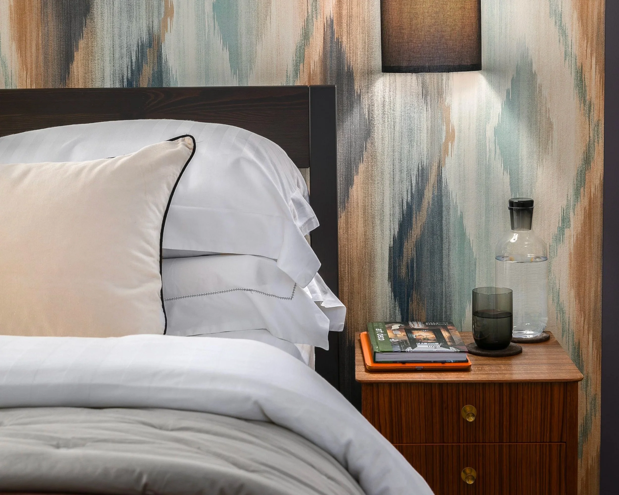 A cozy bedroom scene with a neatly made bed, beige pillow with black piping, and a wooden nightstand holding books, a glass, and a water pitcher with a black lid. The background features a textured, colorful accent wall and a modern wall-mounted lamp