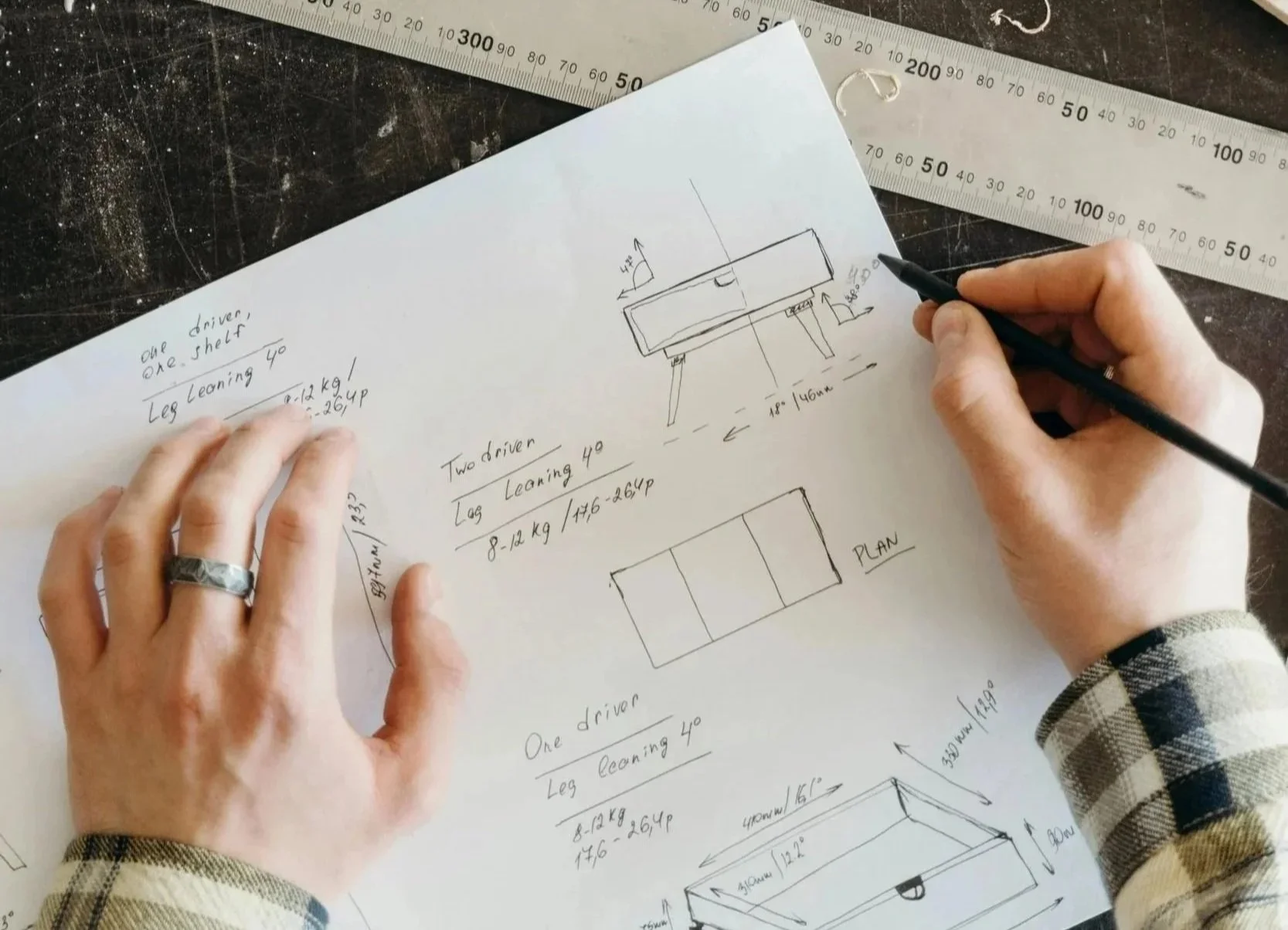 A person sketching design plans on paper with detailed measurements and annotations, with a metal ruler nearby.