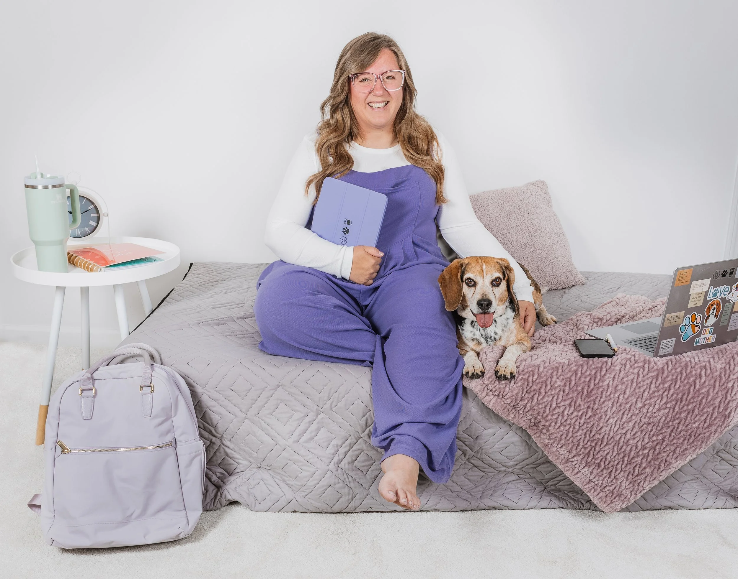 A woman with long wavy hair wearing glasses and purple scrubs sitting on a bed with a dog, holding a laptop. There are a backpack, a laptop, a pink blanket, a cushion, and a backpack nearby, with everyday items on a white side table.