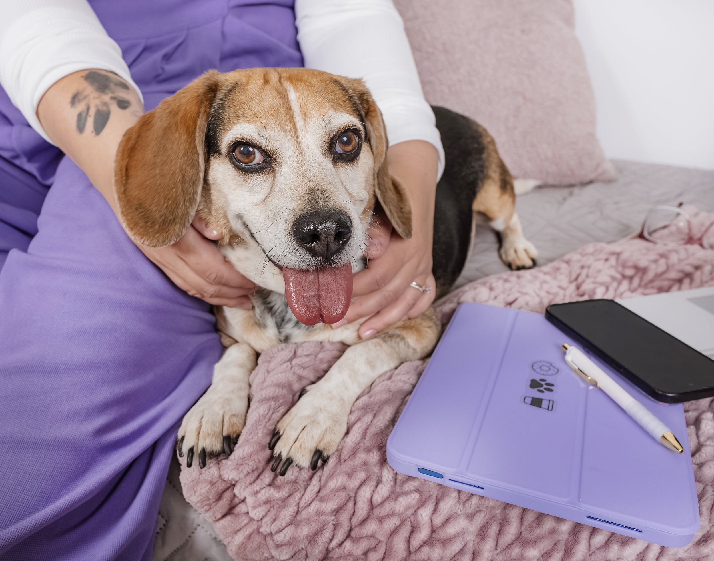 A dog with a tongue out, being held by a person sitting on a bed. The person is wearing purple scrubs and has a tattoo on their arm. On the bed, there is a purple notebook, a pen, and a black phone.