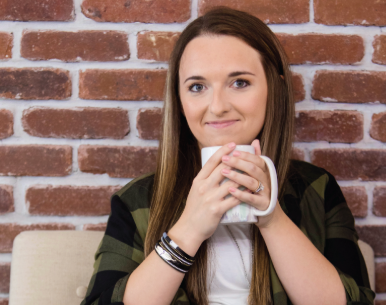Young woman with long brown hair holding a white mug, sitting in front of a brick wall