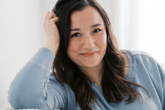 A woman with long dark hair wearing a light blue sweatshirt, smiling and resting her head on her hand