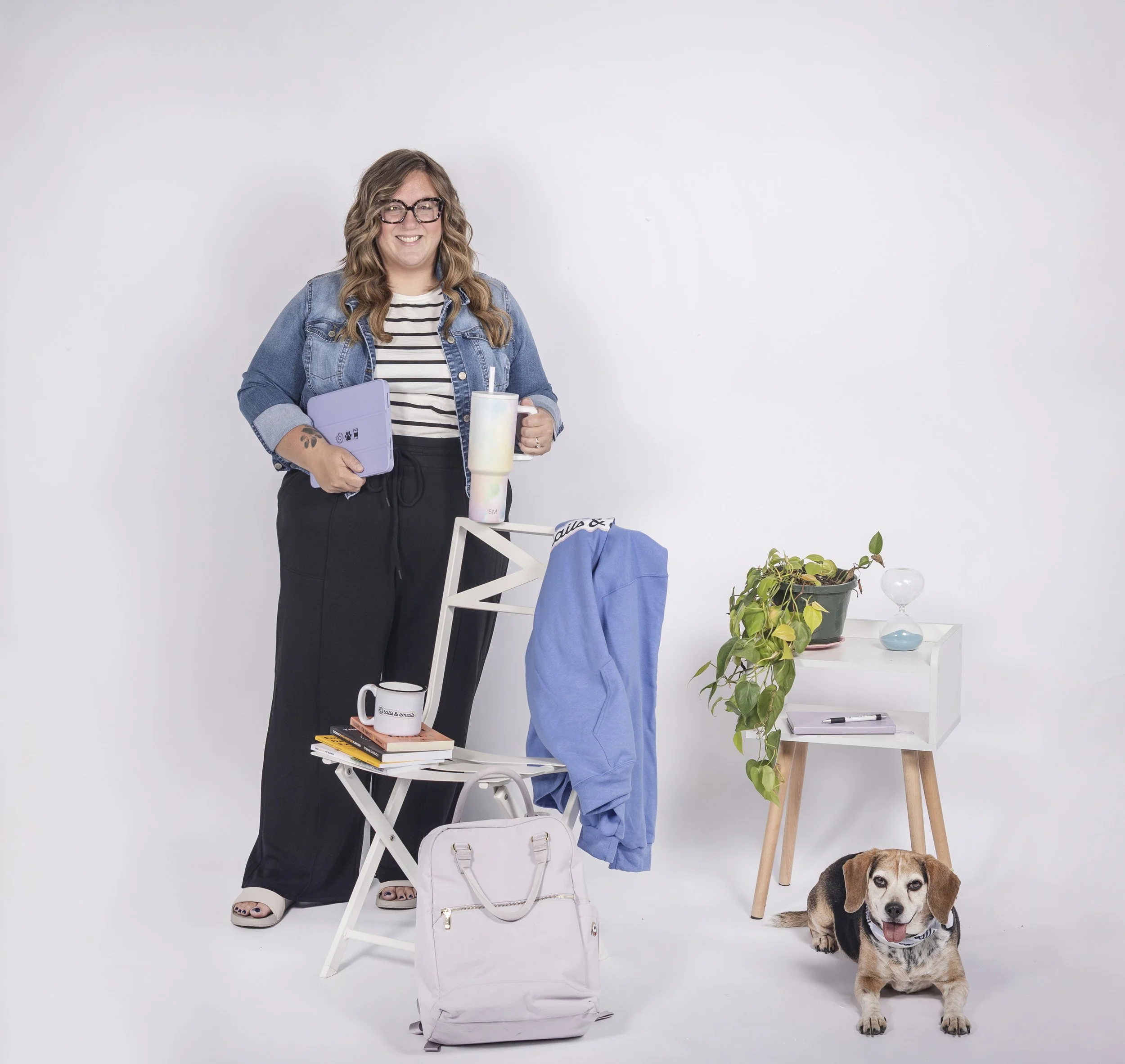 A woman with glasses, a striped shirt, and a denim jacket holding a purple tablet and a large travel mug, standing next to a white chair. There is a stack of books, a white bag, and a white table with a plant and glassware nearby. A dog is sitting on the floor beside her.