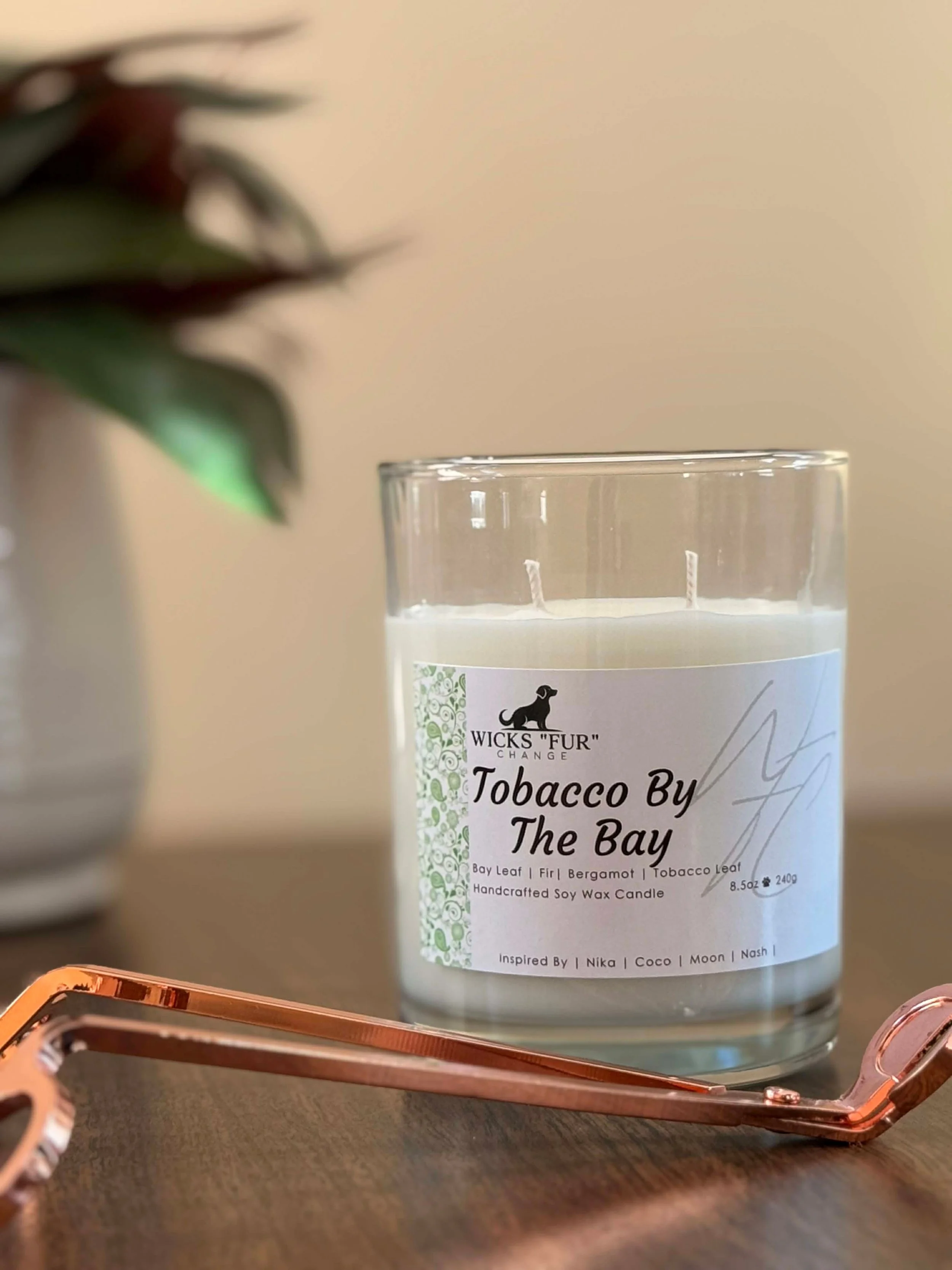 A white scented candle labeled 'Tobacco By The Bay' placed on a metallic stand. The candle is next to a blurred decorative element on the left, with a neutral-colored background.