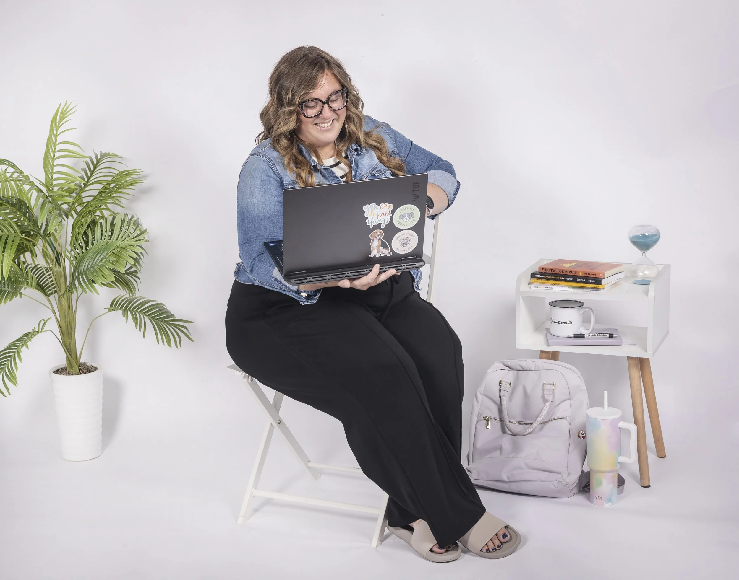 A woman with long wavy hair, glasses, and a denim jacket smiling and looking at a laptop with stickers on it. She is sitting on a white chair in a minimalistic room. To her left, there's a tall potted plant. To her right, a small table with books, a decorative hourglass, a mug, a tumbler, and a backpack.