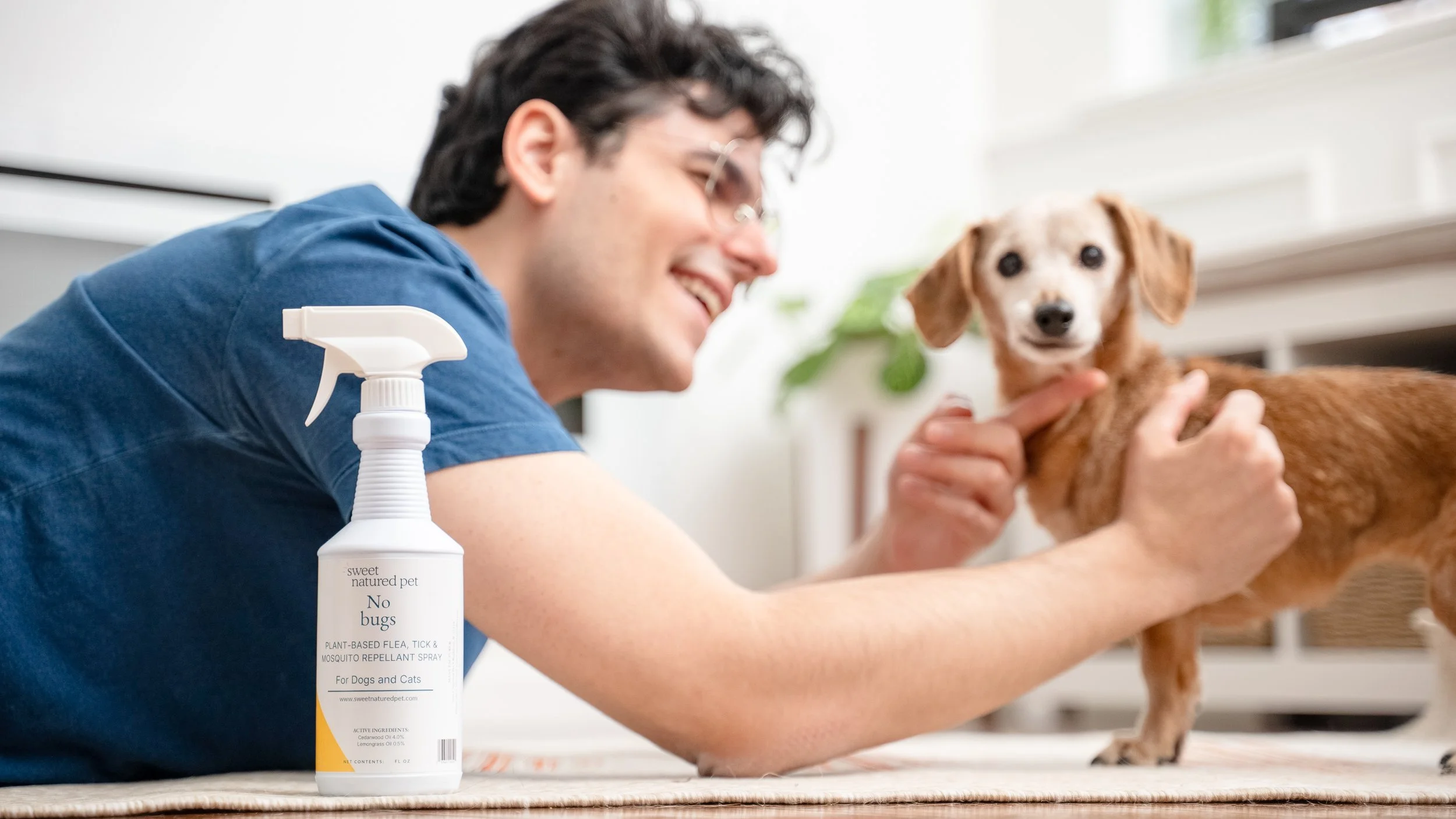 A man smiling while holding a small brown dog, with a spray bottle labeled "No bugs plant-based flea, tick & mosquito repellent spray" in the foreground, in an indoor setting.