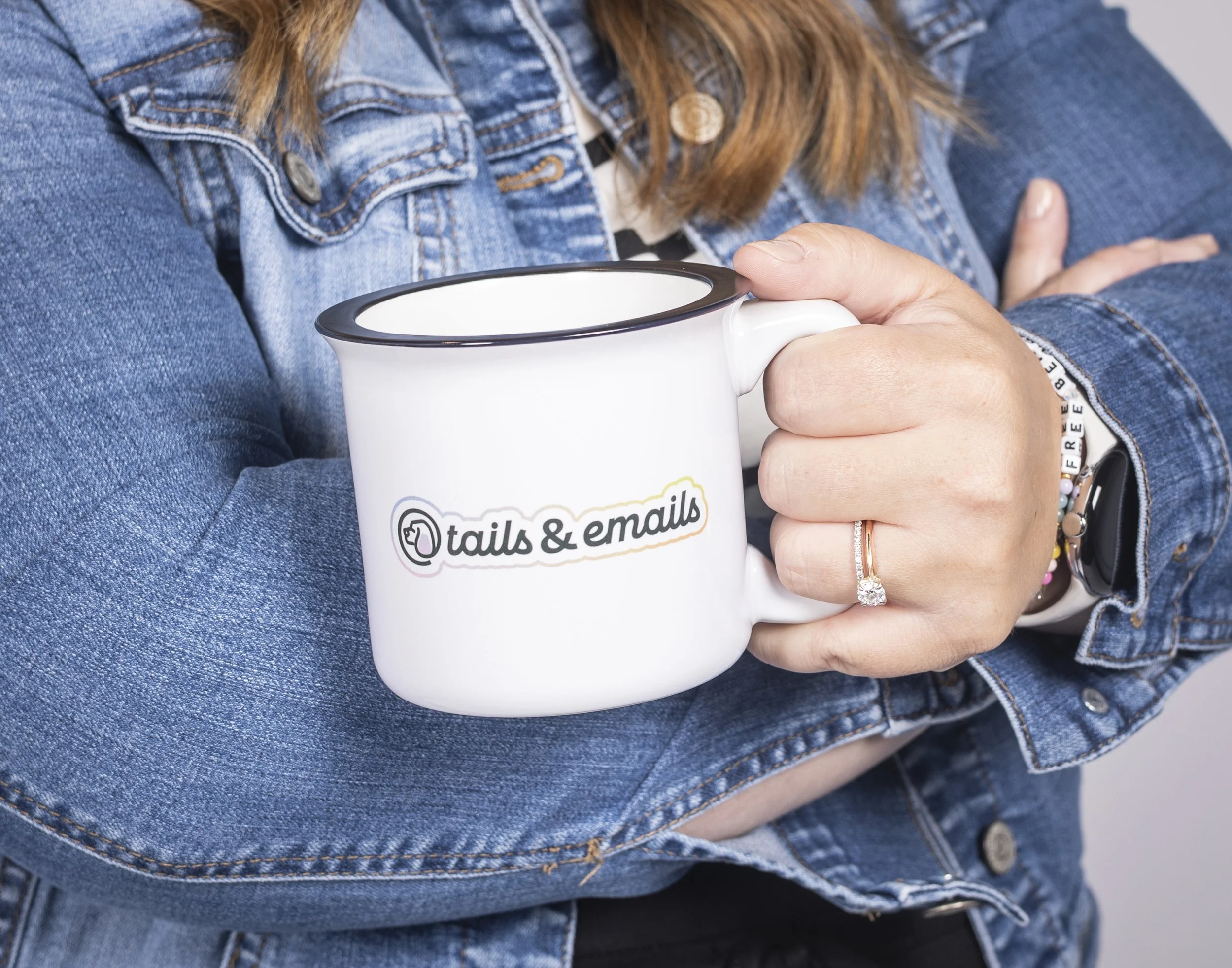 Person wearing a denim jacket and jewelry holding a white mug with text and logo.