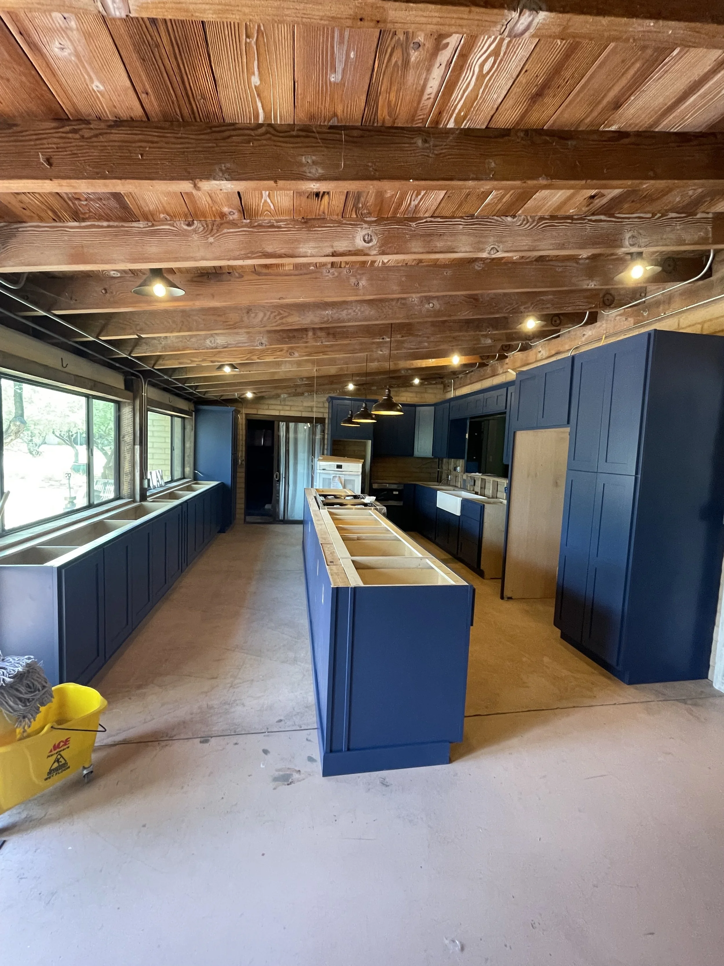 Interior of a kitchen under construction with blue cabinets, a large window, and a wooden ceiling.