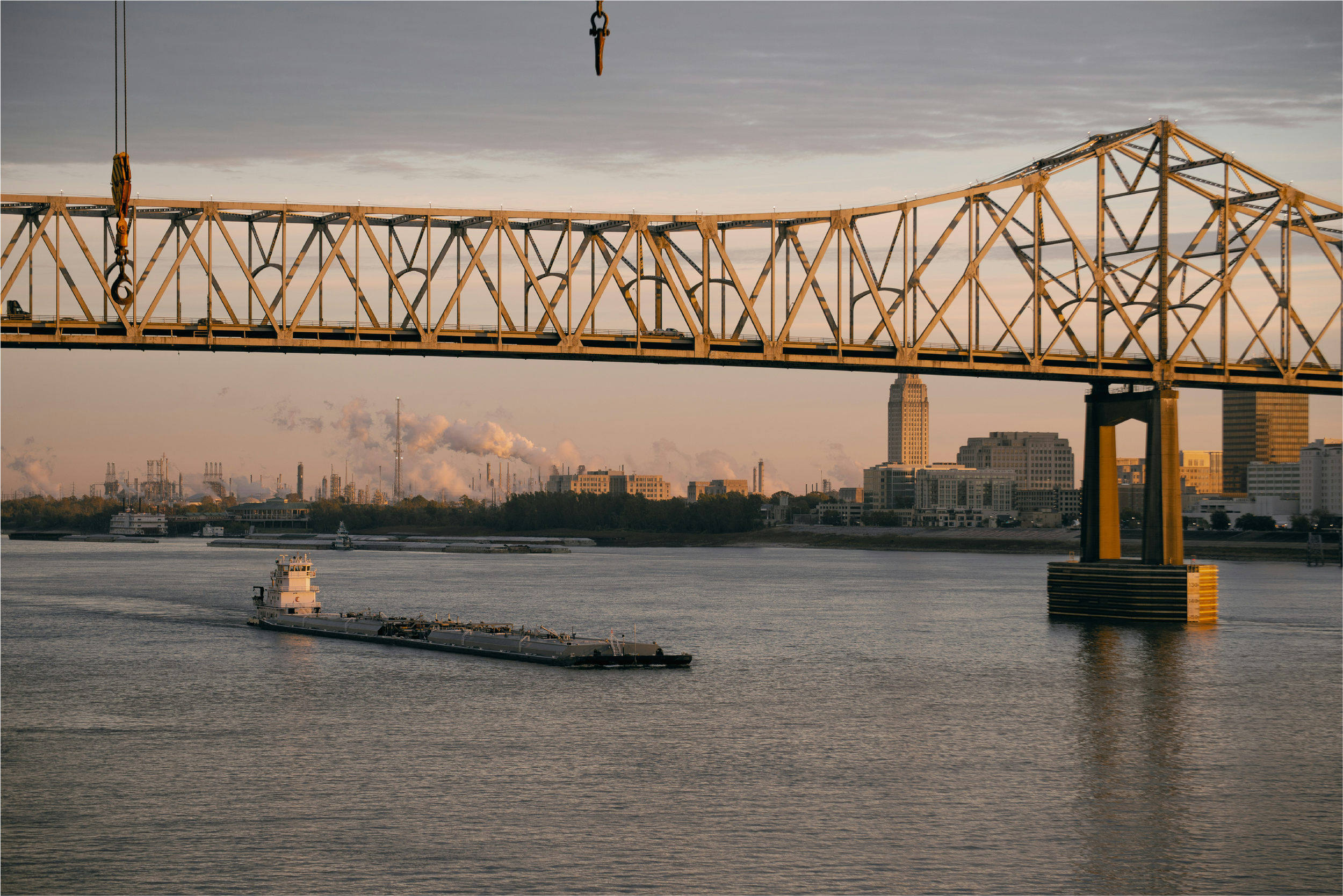 View of a bridge over a river with a barge passing underneath at sunset, city skyline in the background, with clouds and smoke.