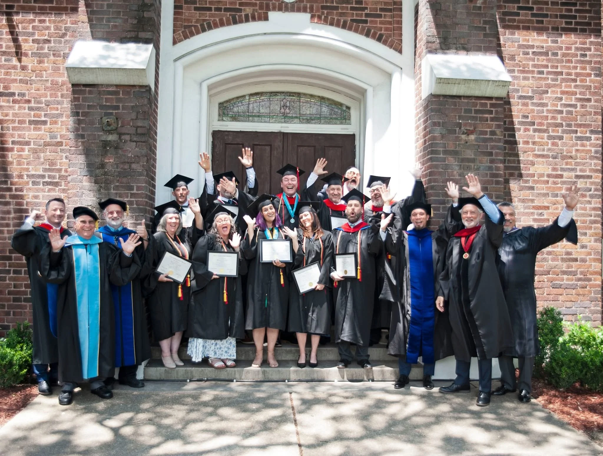 A group of jubilant graduates in black robes and caps, some holding diplomas, standing on steps in front of a brick building, celebrating their graduation on a sunny day.