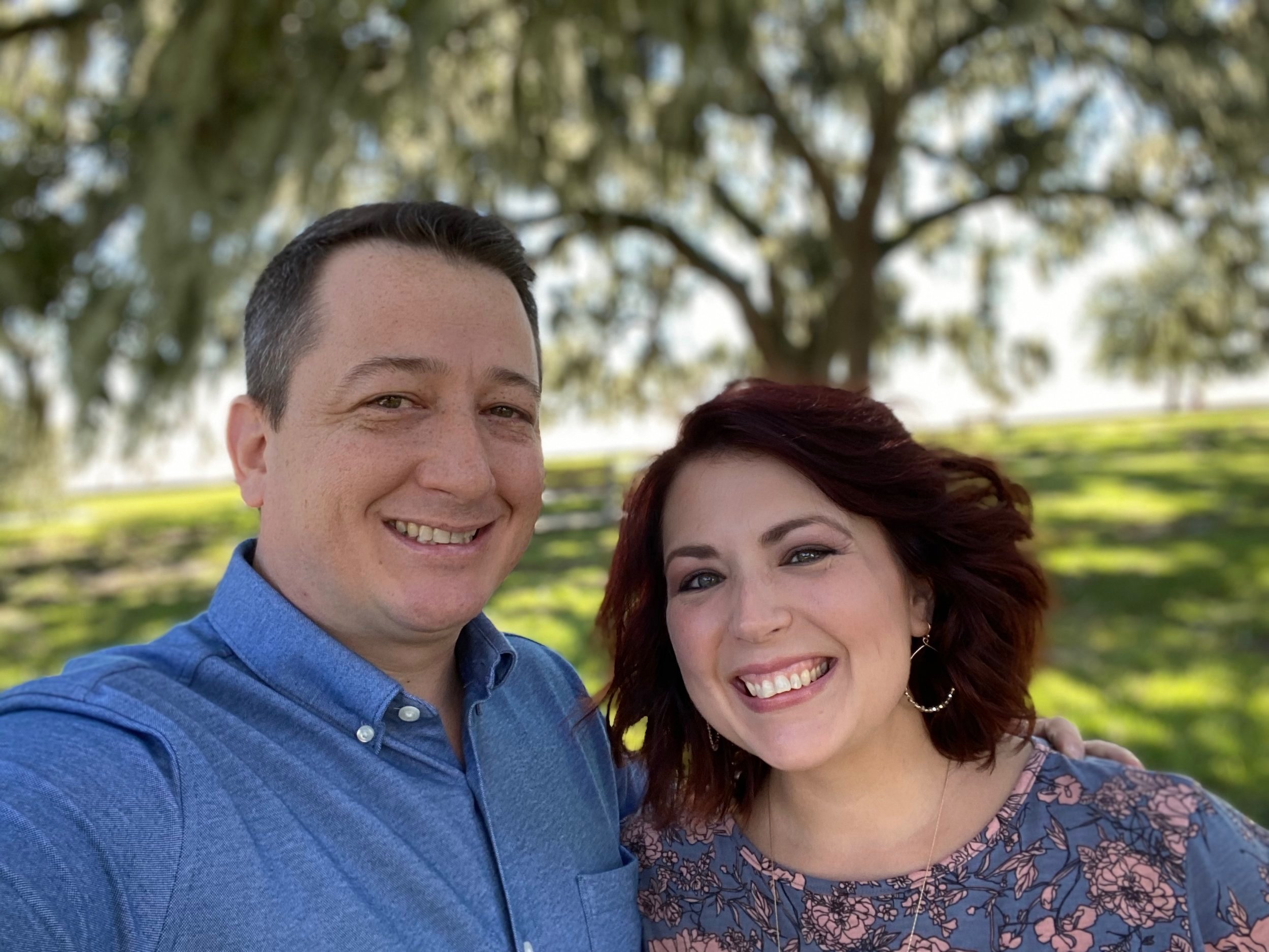 Pastors Al & Jenn smiling outdoors, with trees and grass in the background.
