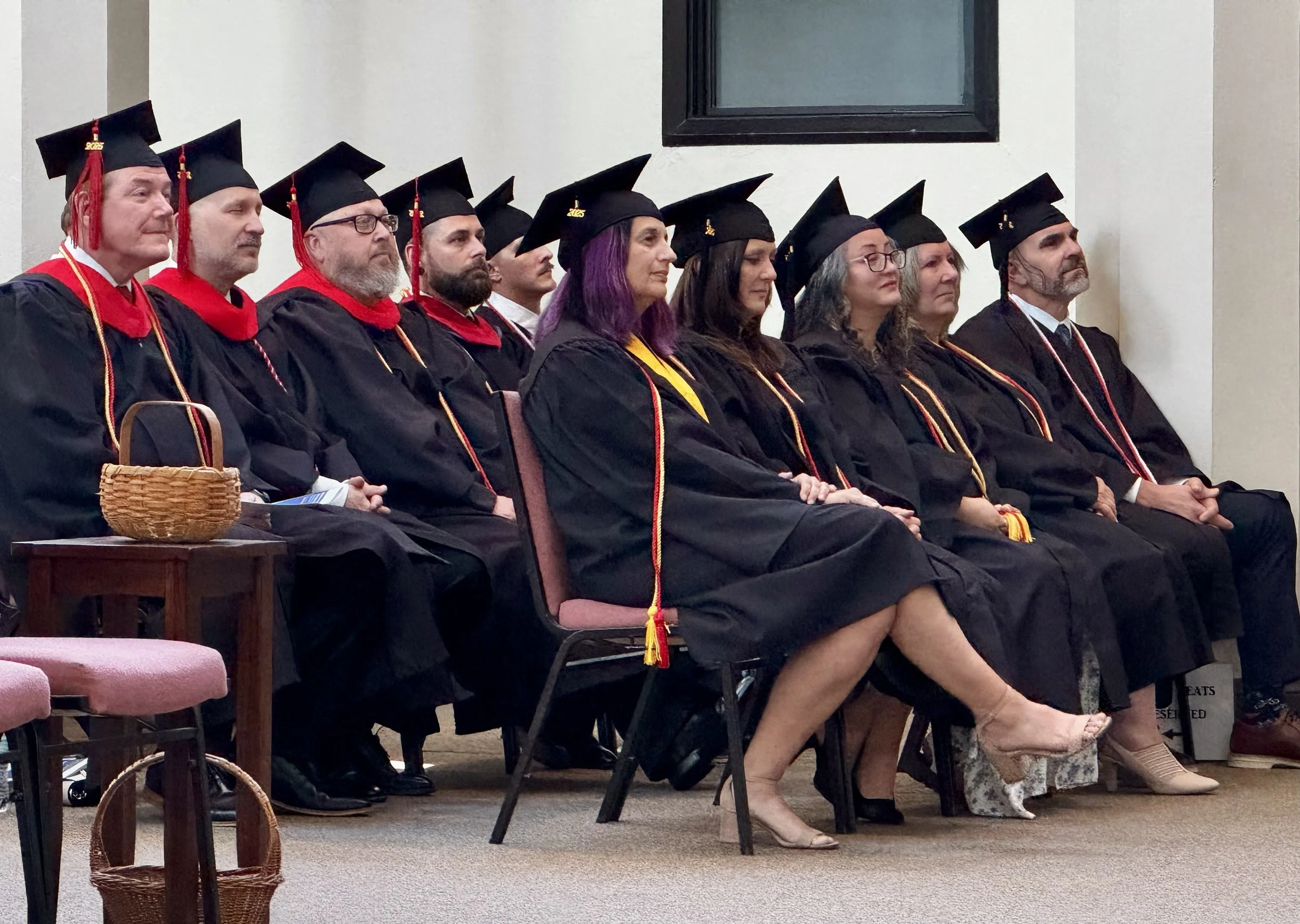 Group of graduates in black caps and gowns sitting during a ceremony, some with cords and stoles, in an indoor setting with a white wall and window behind them.