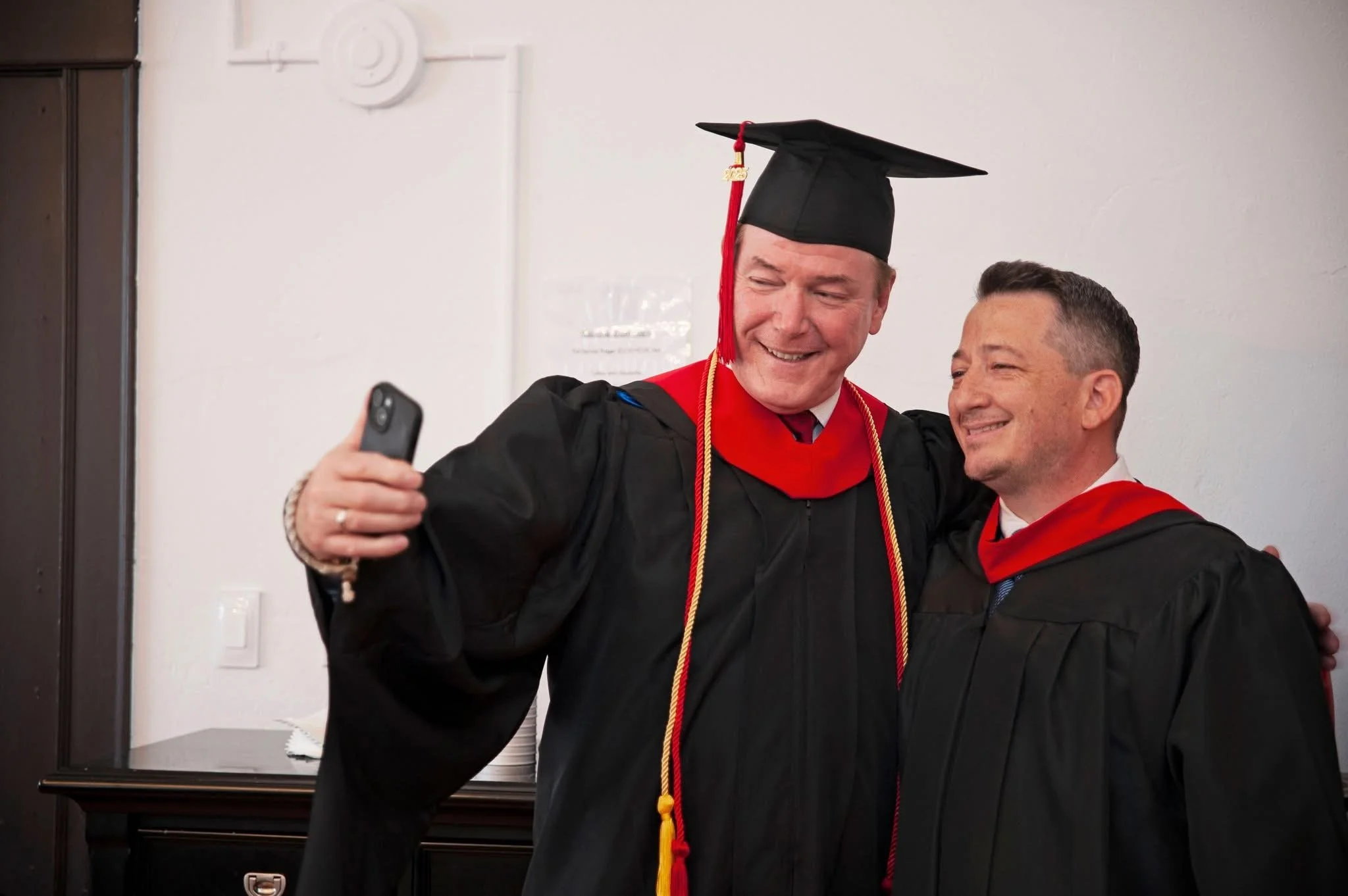 Two men in caps and gowns, taking a selfie at graduation ceremony.