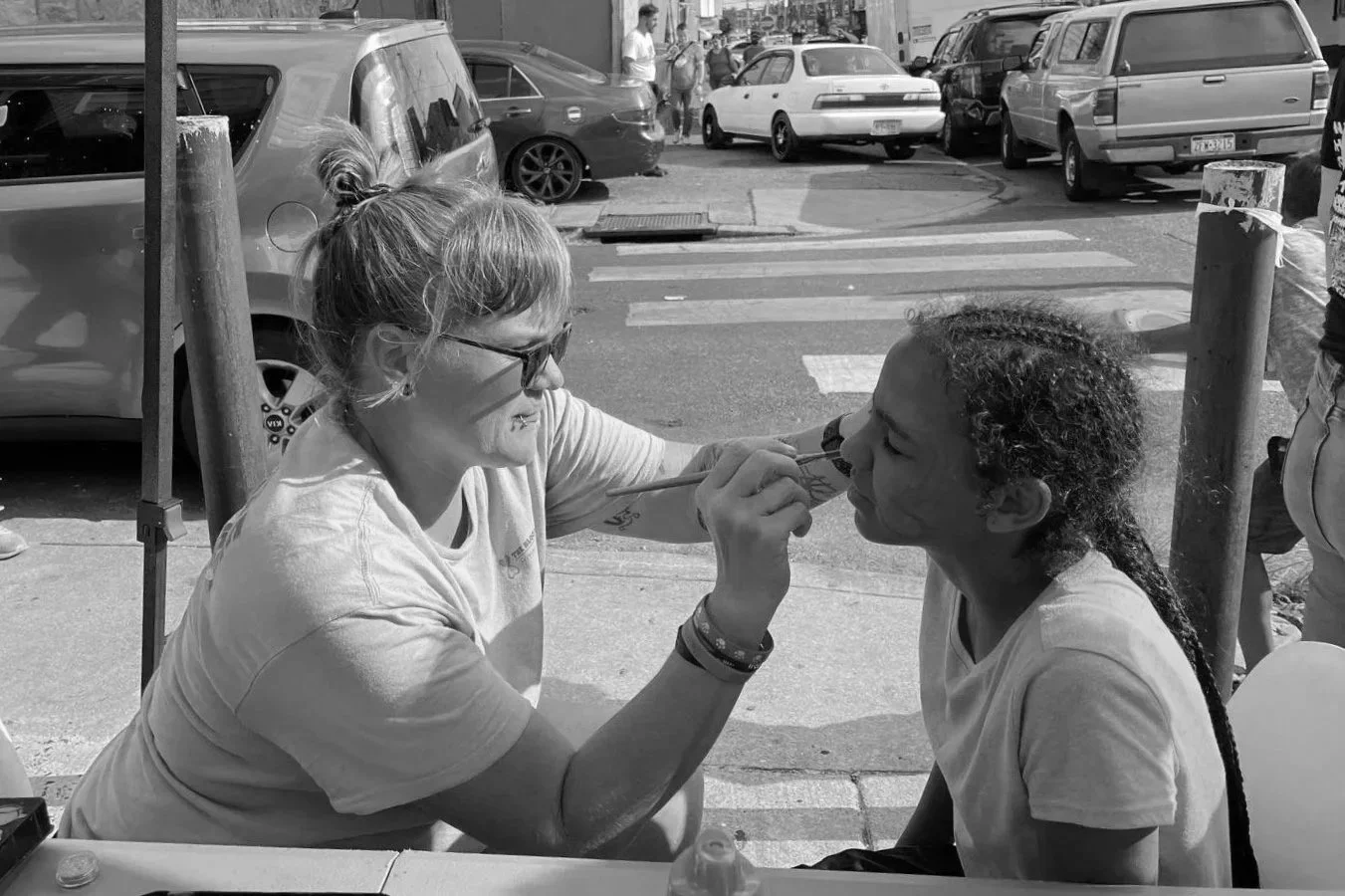 A woman painting a young girl’s face outdoors on a city sidewalk, with cars and pedestrians in the background.
