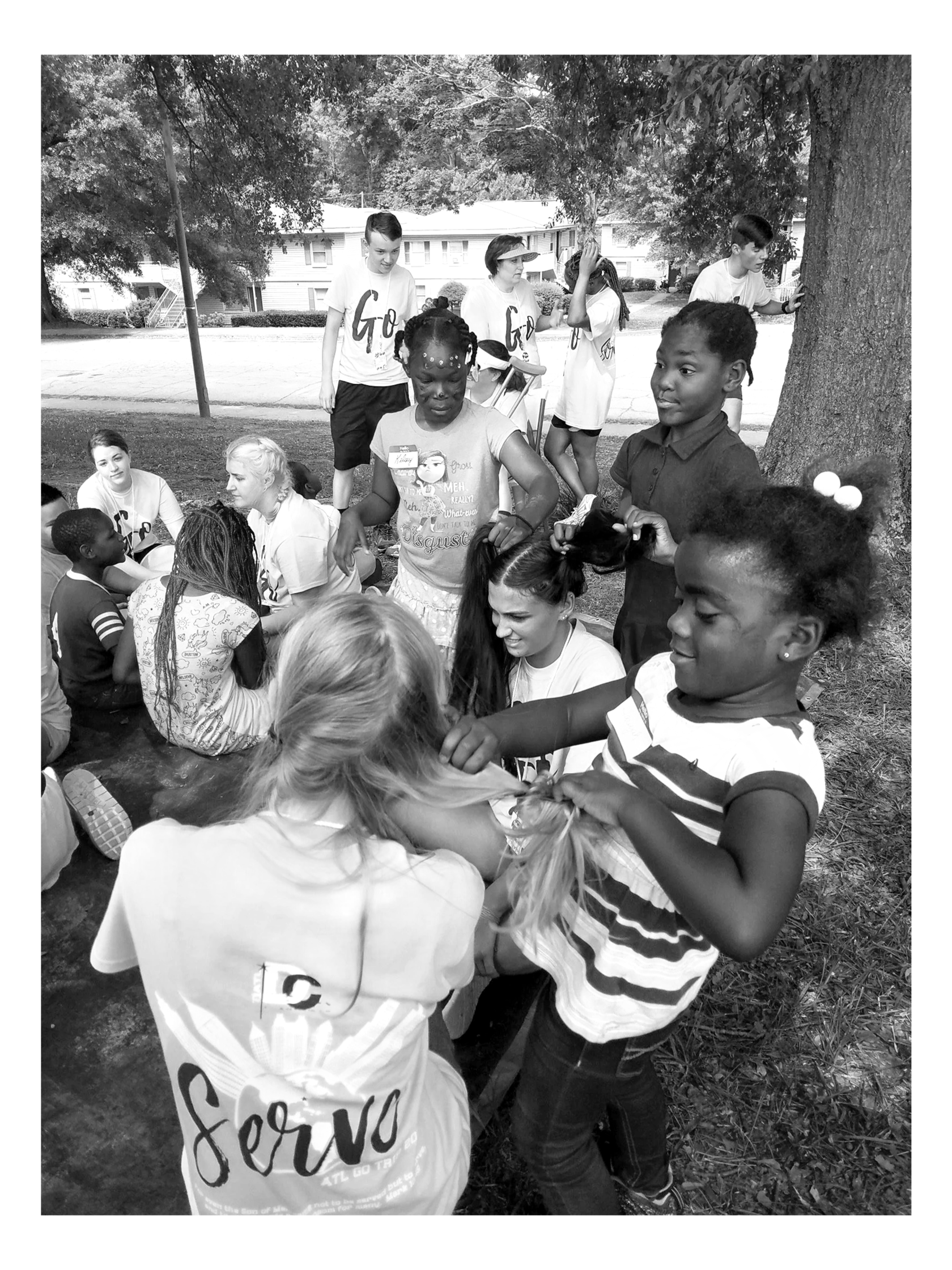 Children and teenagers gathered outdoors under a large tree, with some sitting on the grass and others standing, engaging in a communal activity.