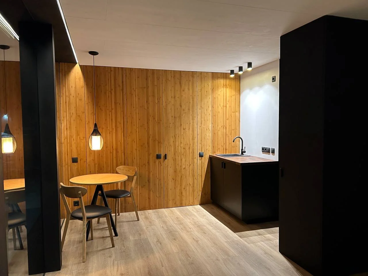 Modern minimalist kitchen with wood-paneled wall, black cabinets, small dining table with two chairs, pendant lighting, and a sink.