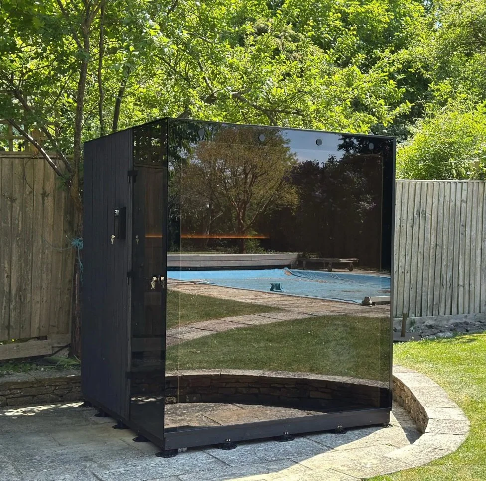 A black outdoor cabinet with a glass front, placed on a stone patio in a backyard. The backyard has a pool covered with a blue tarp, surrounded by a grassy area and trees, with a wooden fence in the background.