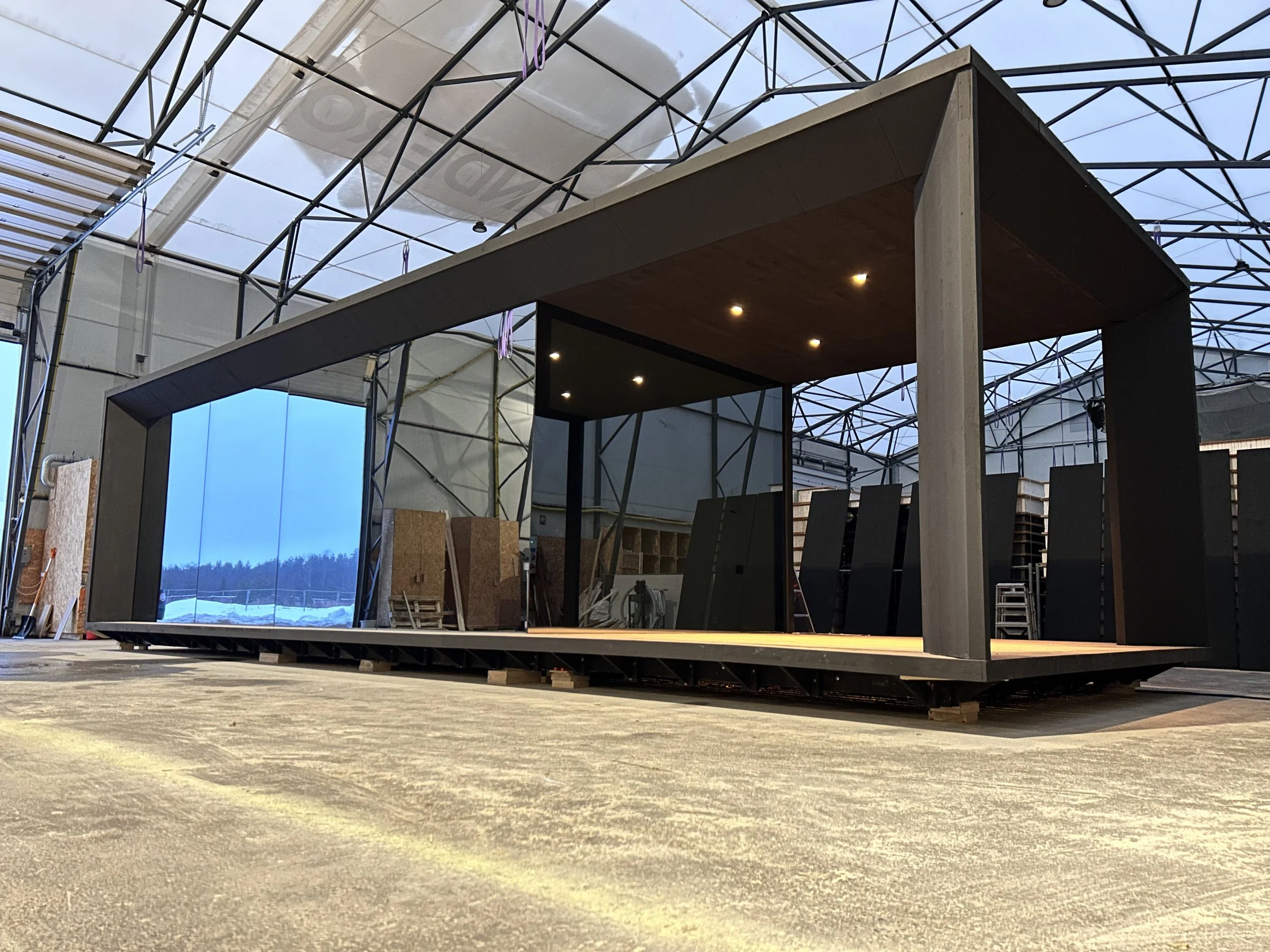 A modern, minimalist indoor stage or pavilion with a black frame and wooden ceiling, under construction inside a large industrial building with a glass ceiling and metal framework.