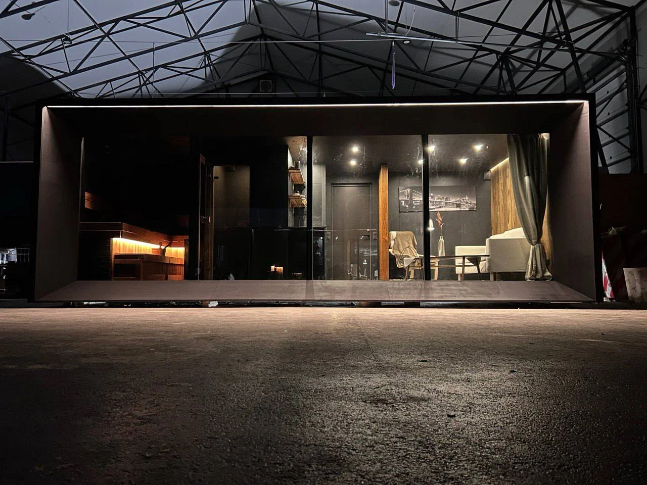 Modern tiny house or office space with glass front wall, interior lighting, sofa, chair, and decorative elements, set inside a larger industrial structure.
