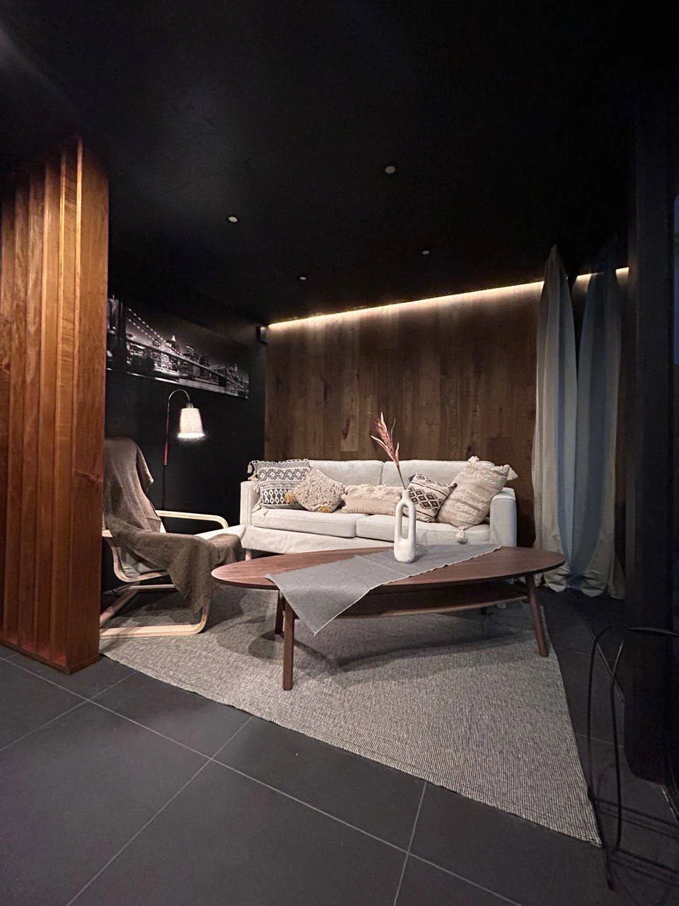Living room with white sofa, wooden coffee table with a beige runner, armchair with a blanket, a floor lamp, and wooden panel accents on the walls, with curtains and dim lighting.