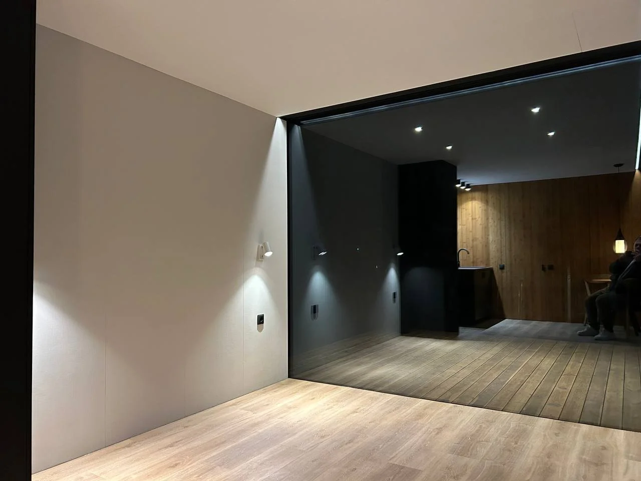 Modern interior room with wooden flooring, beige and dark walls, and recessed ceiling lights; a person sitting on a chair is visible on the right side.