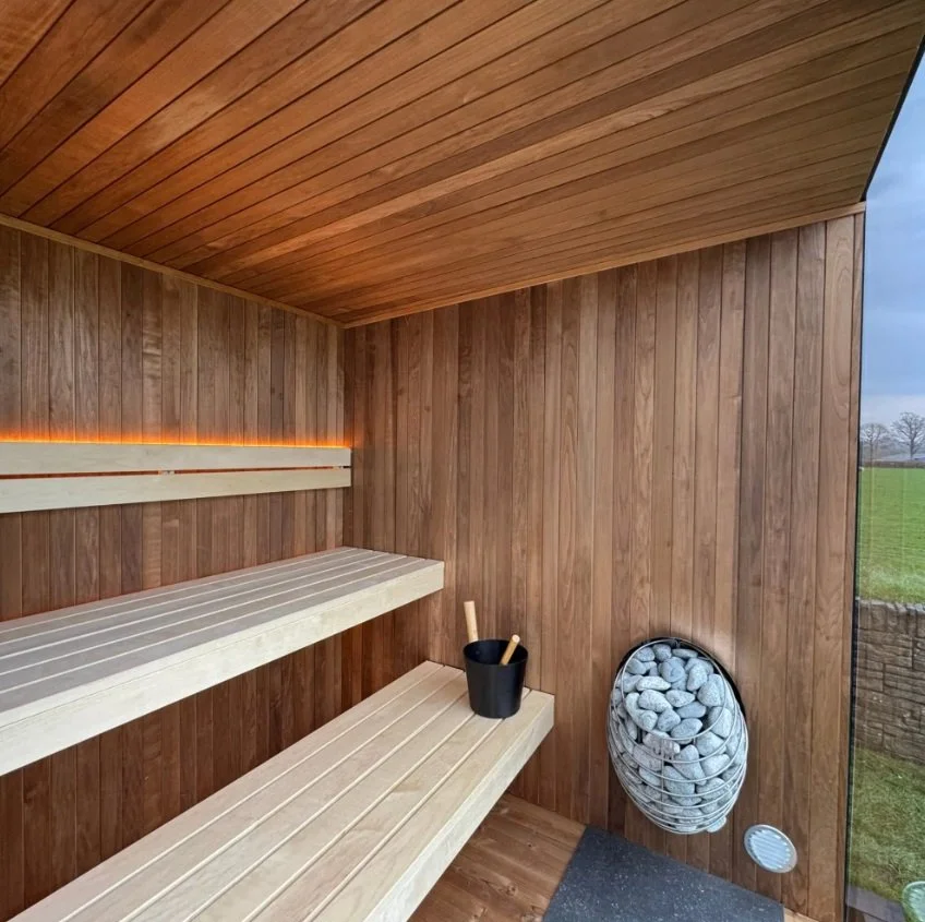 Interior of a modern sauna with wooden walls, benches, a bucket with ladles, and a heater filled with stones. For full retreat and energy regeneration.