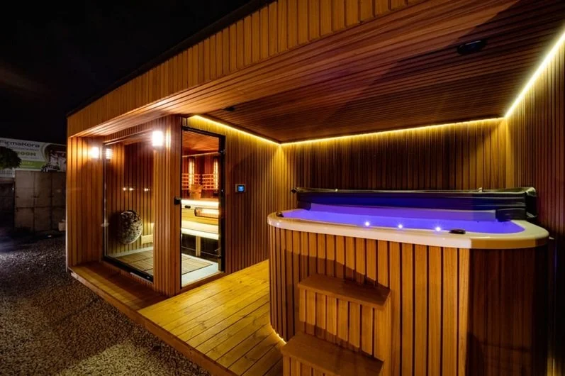 Outdoor hot tub with LED lighting and a sauna or steam room with glass door, all surrounded by wooden paneling.