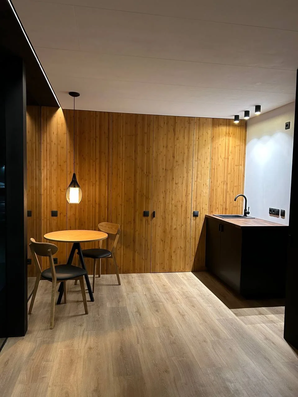 Modern kitchenette with wooden wall panels, a small round table with two chairs, a hanging pendant light, black cabinetry with a sink, and recessed ceiling lights.