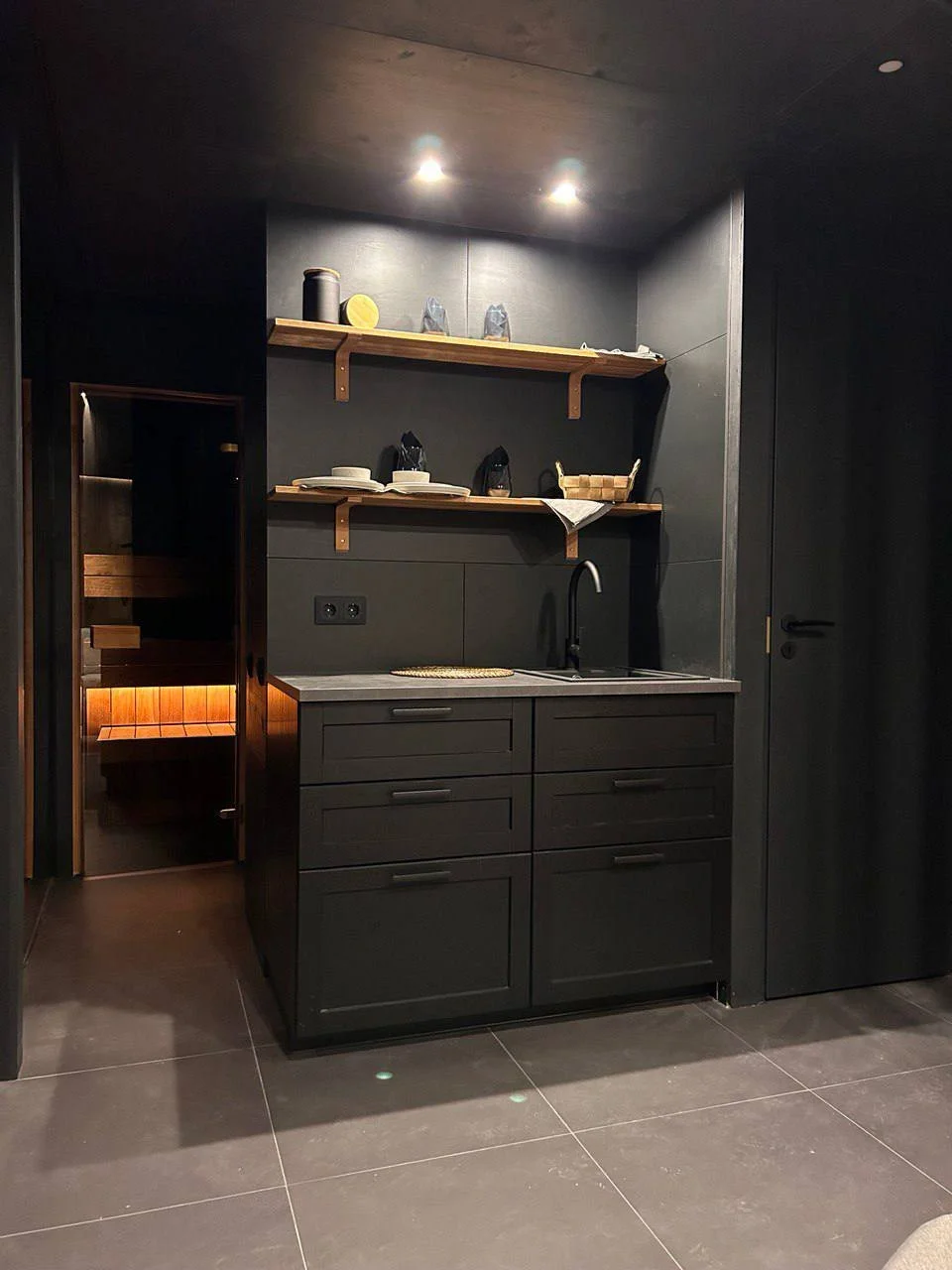 A small kitchen area with black cabinets, a grey countertop, and two wooden shelves holding various dishes and containers, with a black sink and faucet. There is a door to a sauna or steam room to the left and a black door to the right. The walls, ce