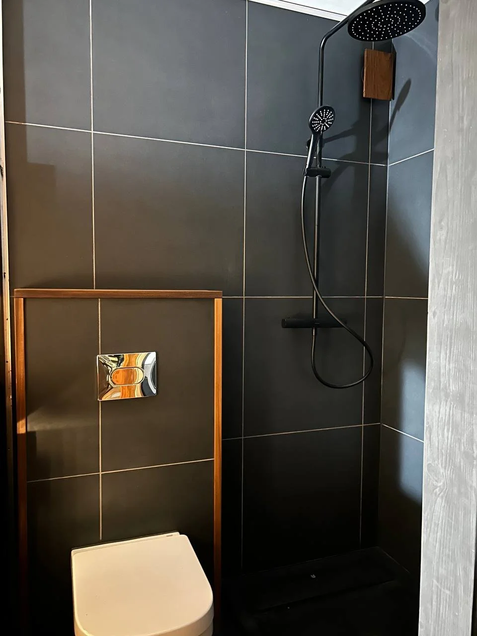 A modern bathroom shower area with dark gray tiles, a black rainfall showerhead, and a wooden accent wall.