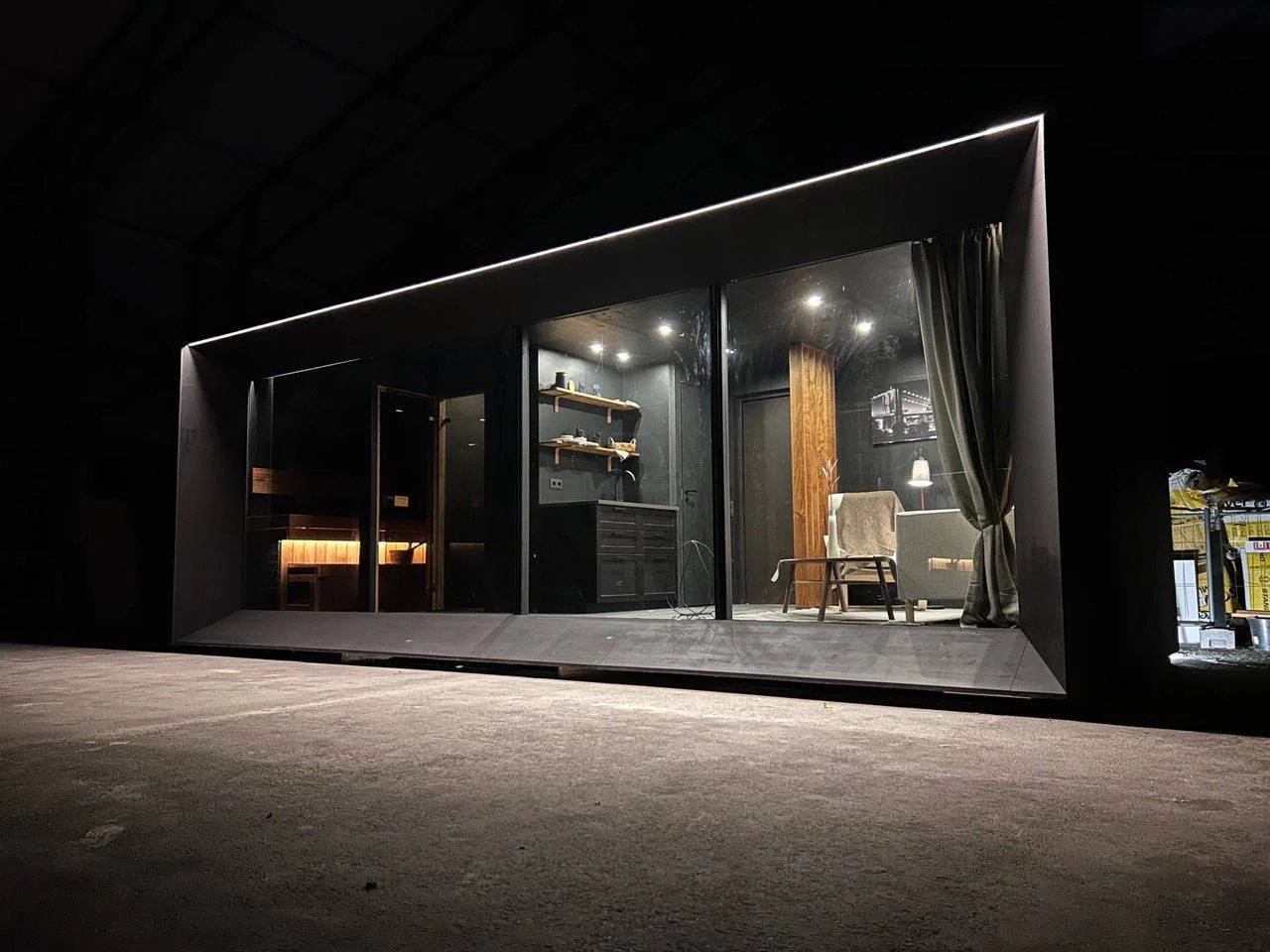 Night view of a modern building with large glass windows revealing a cozy living space inside, including a chair, table lamp, and shelves.