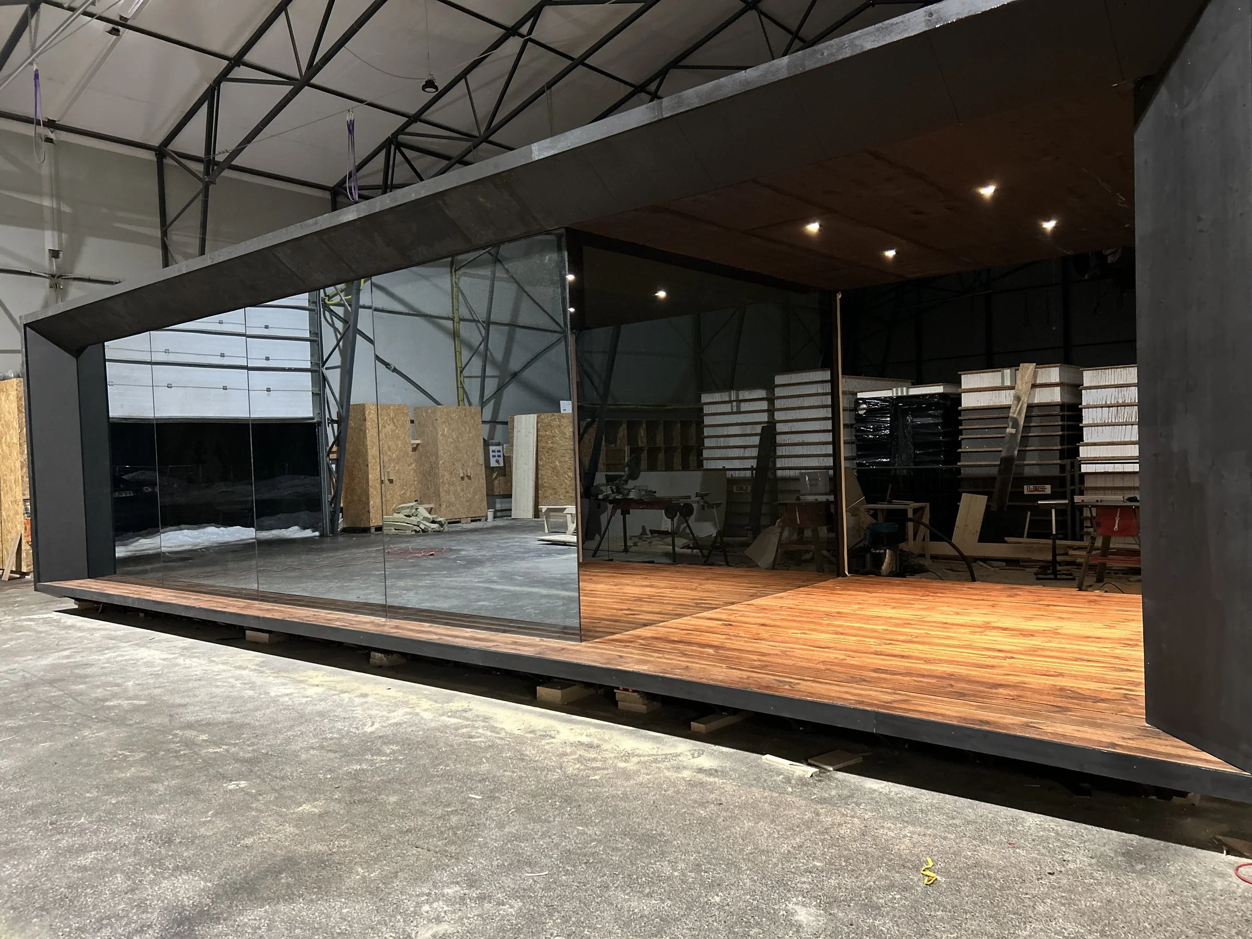 Interior view of a modern, unfinished building with a large glass wall and a wooden floor, surrounded by construction materials and equipment.