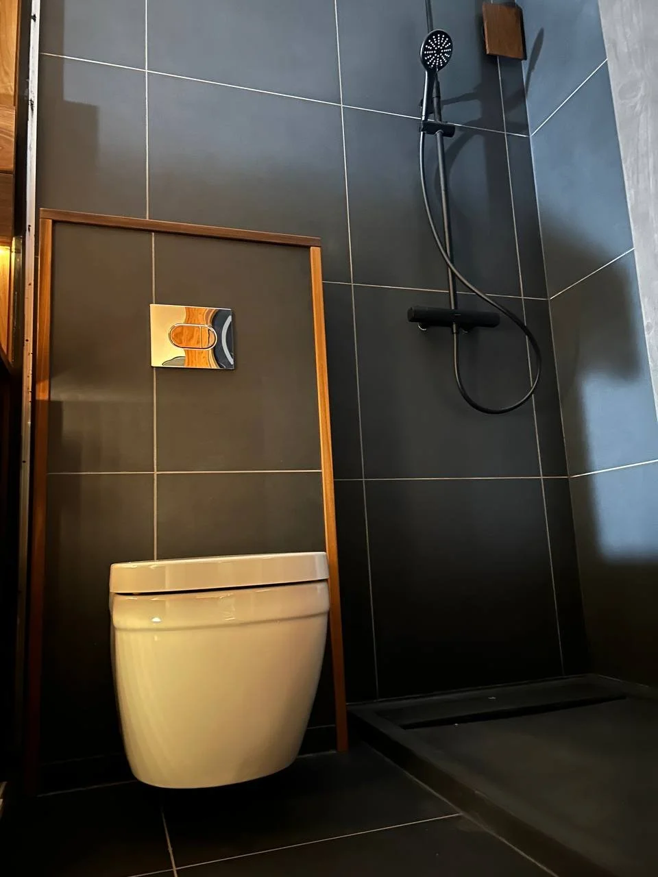 Modern bathroom with dark gray tiles, a wall-mounted toilet, and a walk-in shower with a black showerhead and hand-held shower wand.